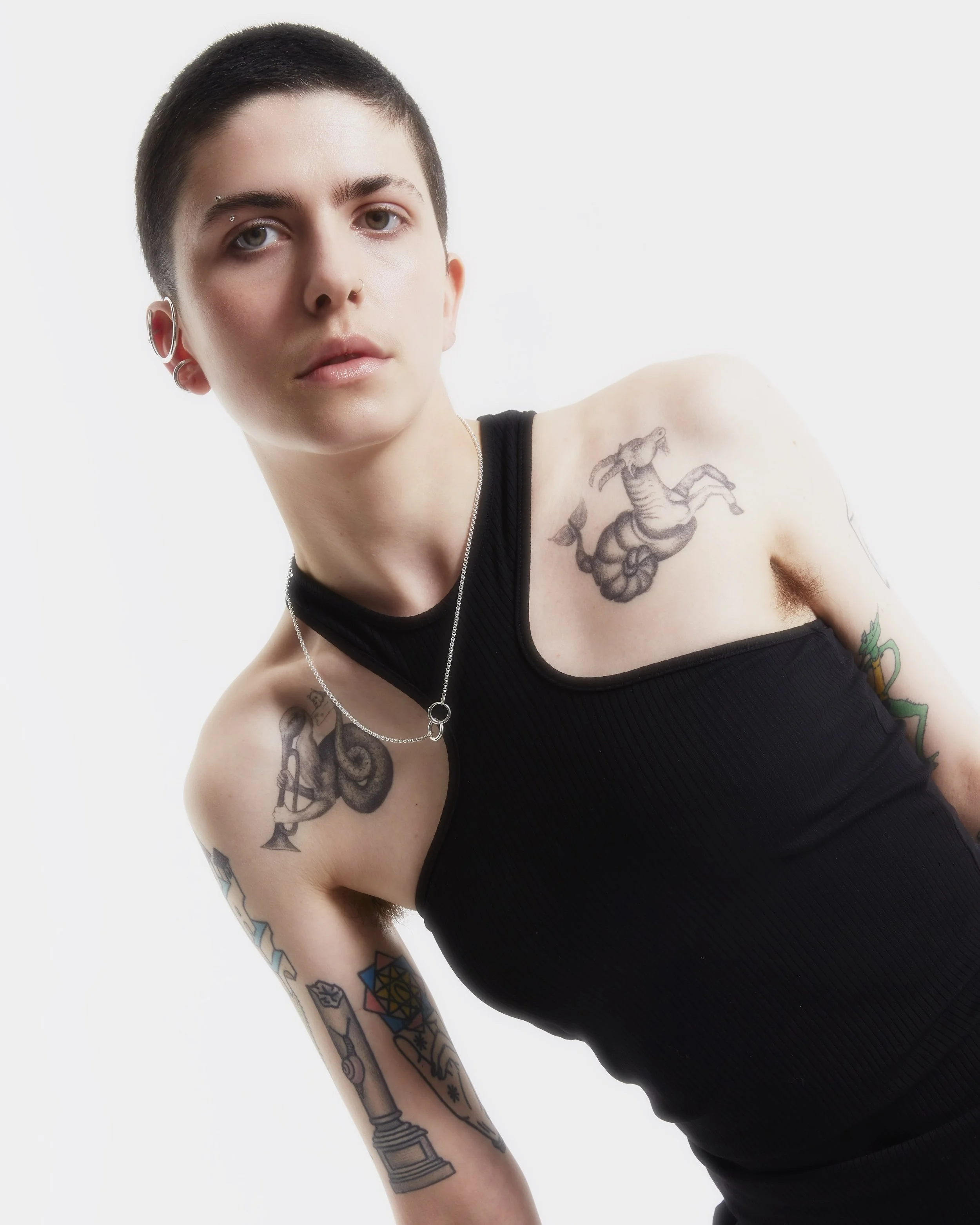 Young person with short dark hair, multiple tattoos on arms and shoulder, wearing a black sleeveless top, posing against a plain white background.
