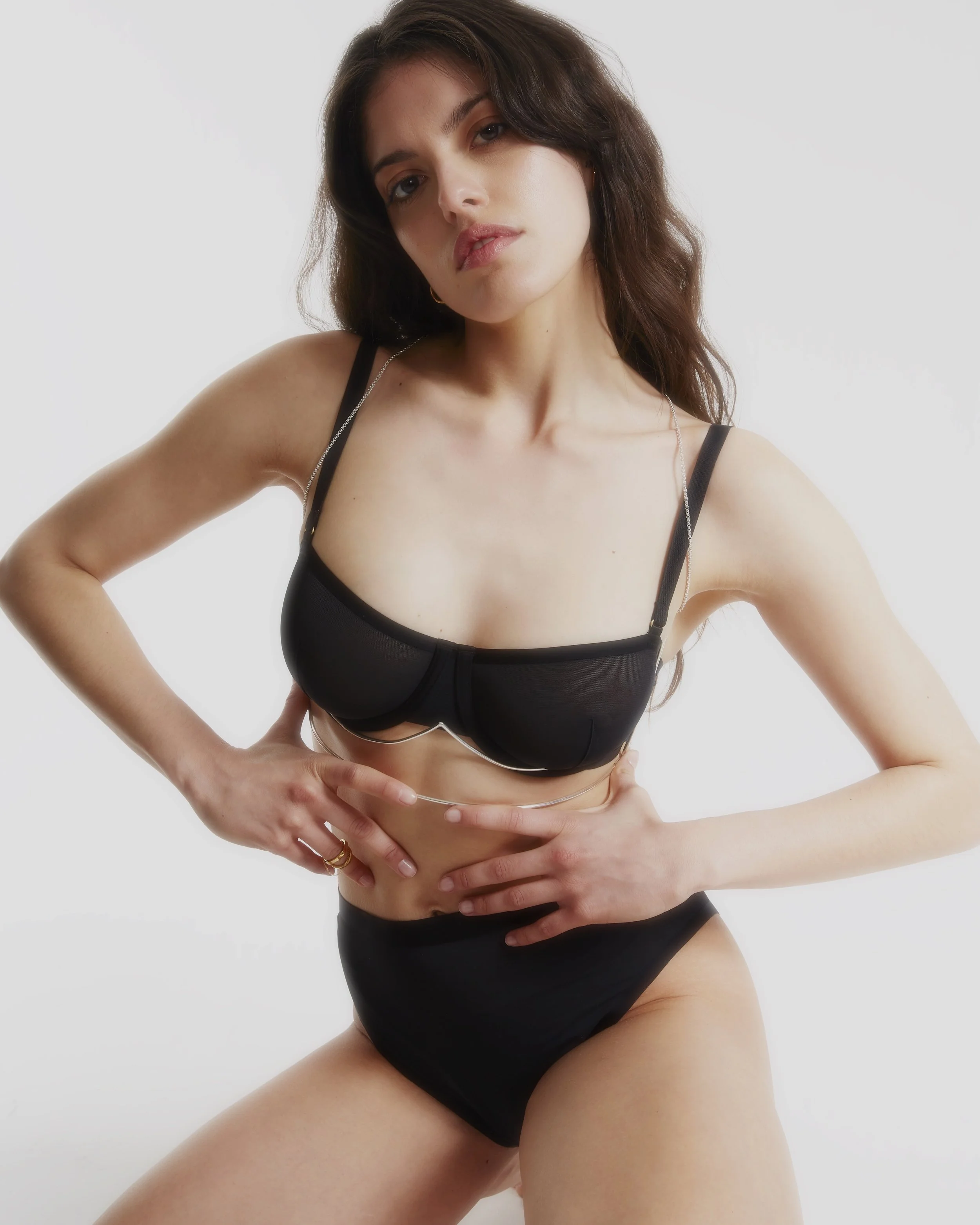 Woman with long brown hair posing in black lingerie against a white background.
