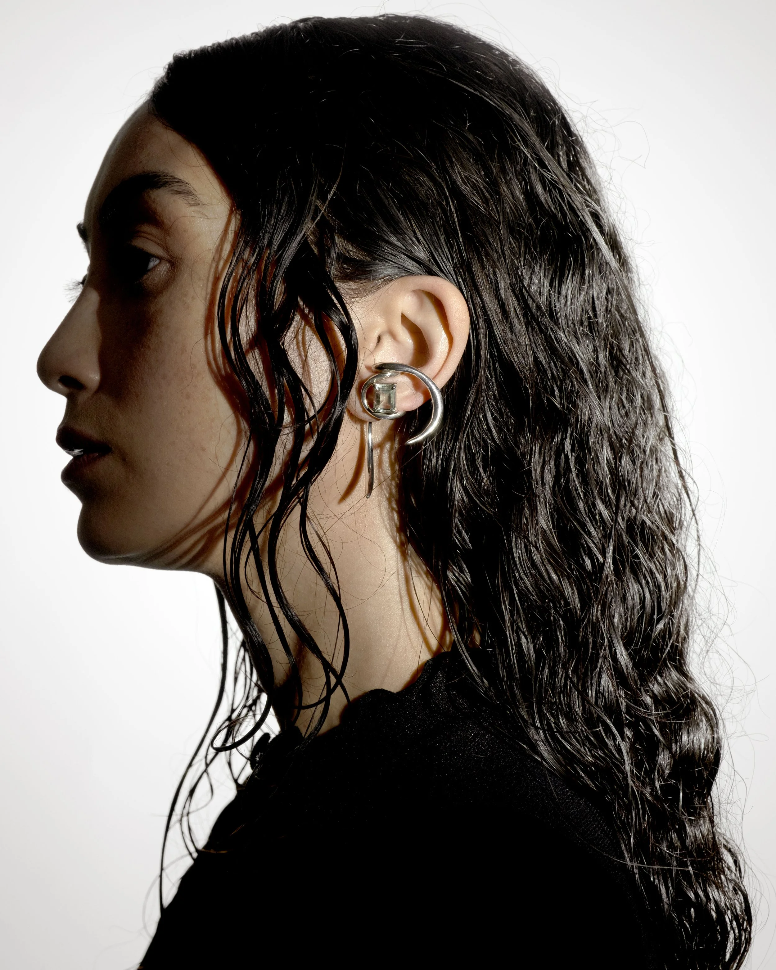 Side profile of a young woman with wet, wavy dark hair, wearing large silver earrings and a black top against a plain white background.