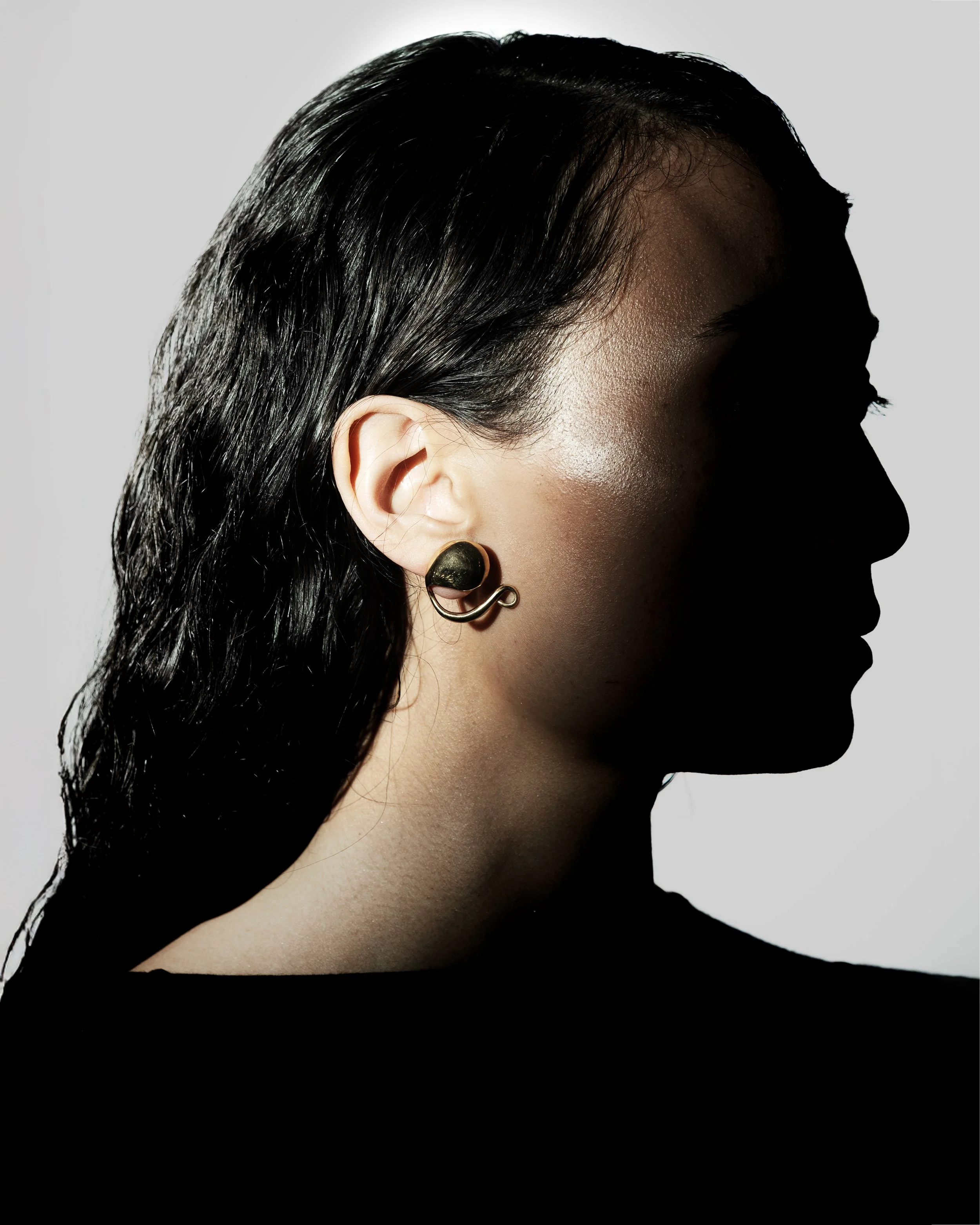 Side profile of a woman with dark, wet hair wearing a black earring, with her face mostly in shadow.