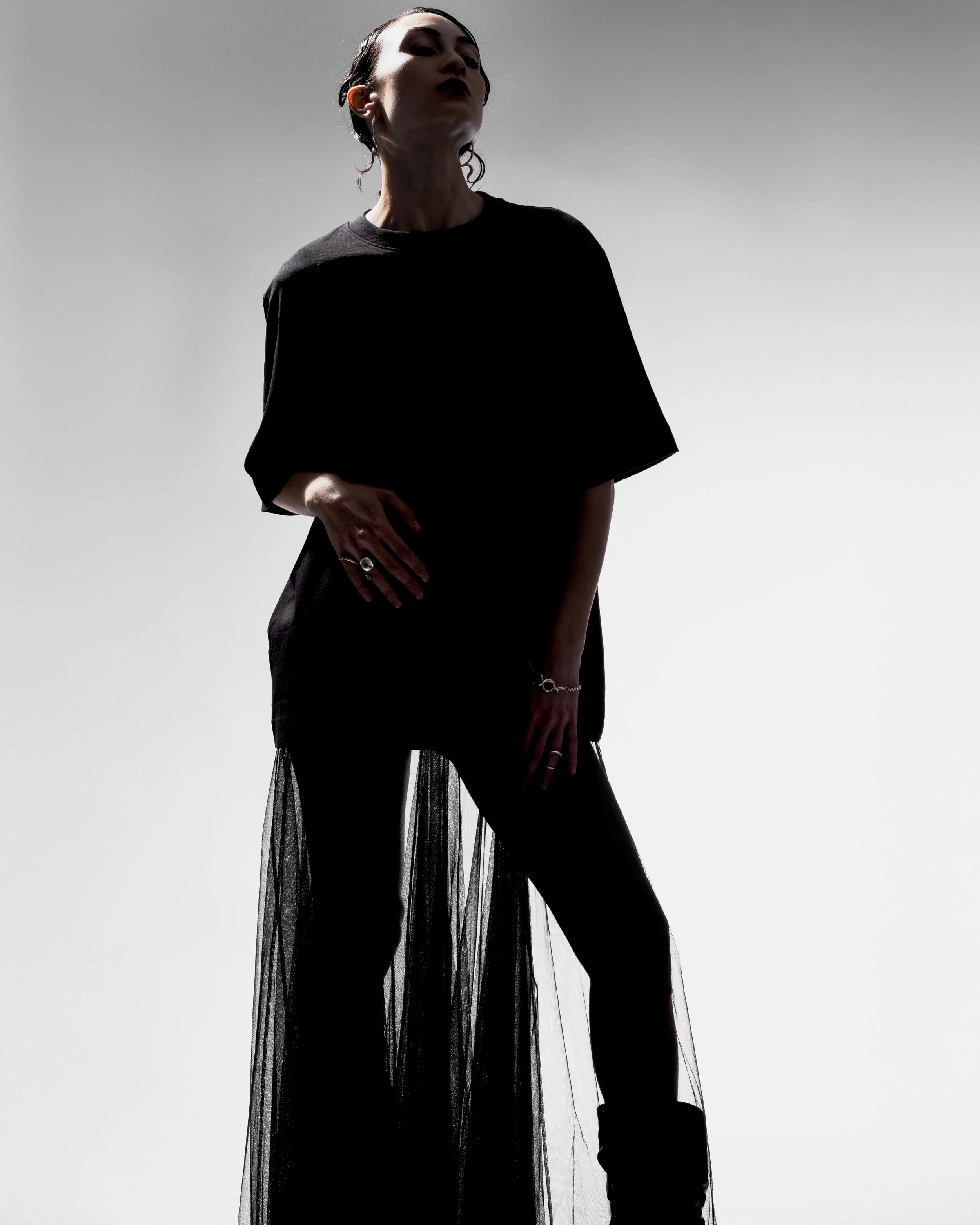A woman in black clothing, standing against a gray background, with her head slightly tilted and her face partially obscured by shadows.