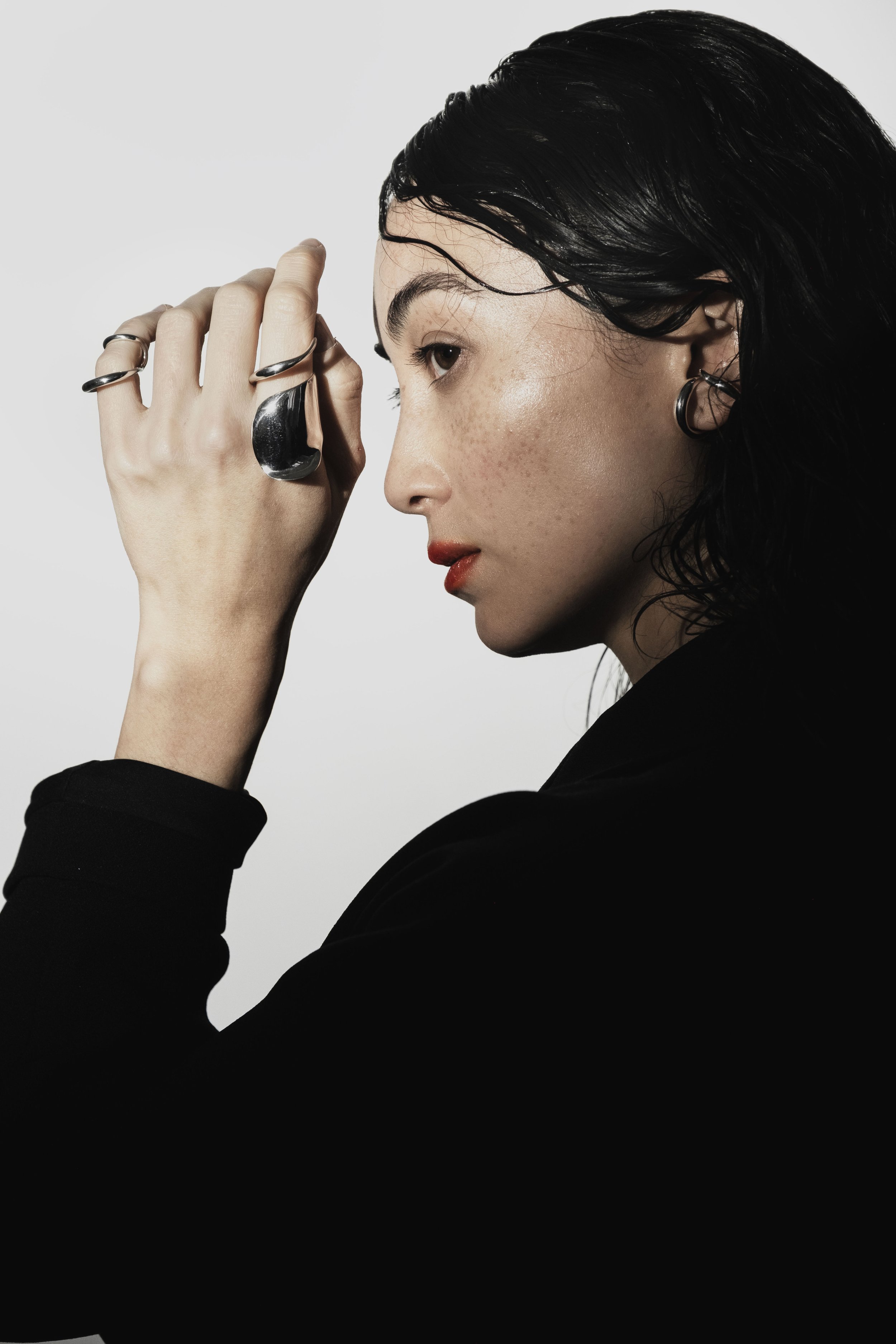 A woman with black hair and red lipstick, wearing jewelry, shown in profile with her right hand near her face, against a plain background.
