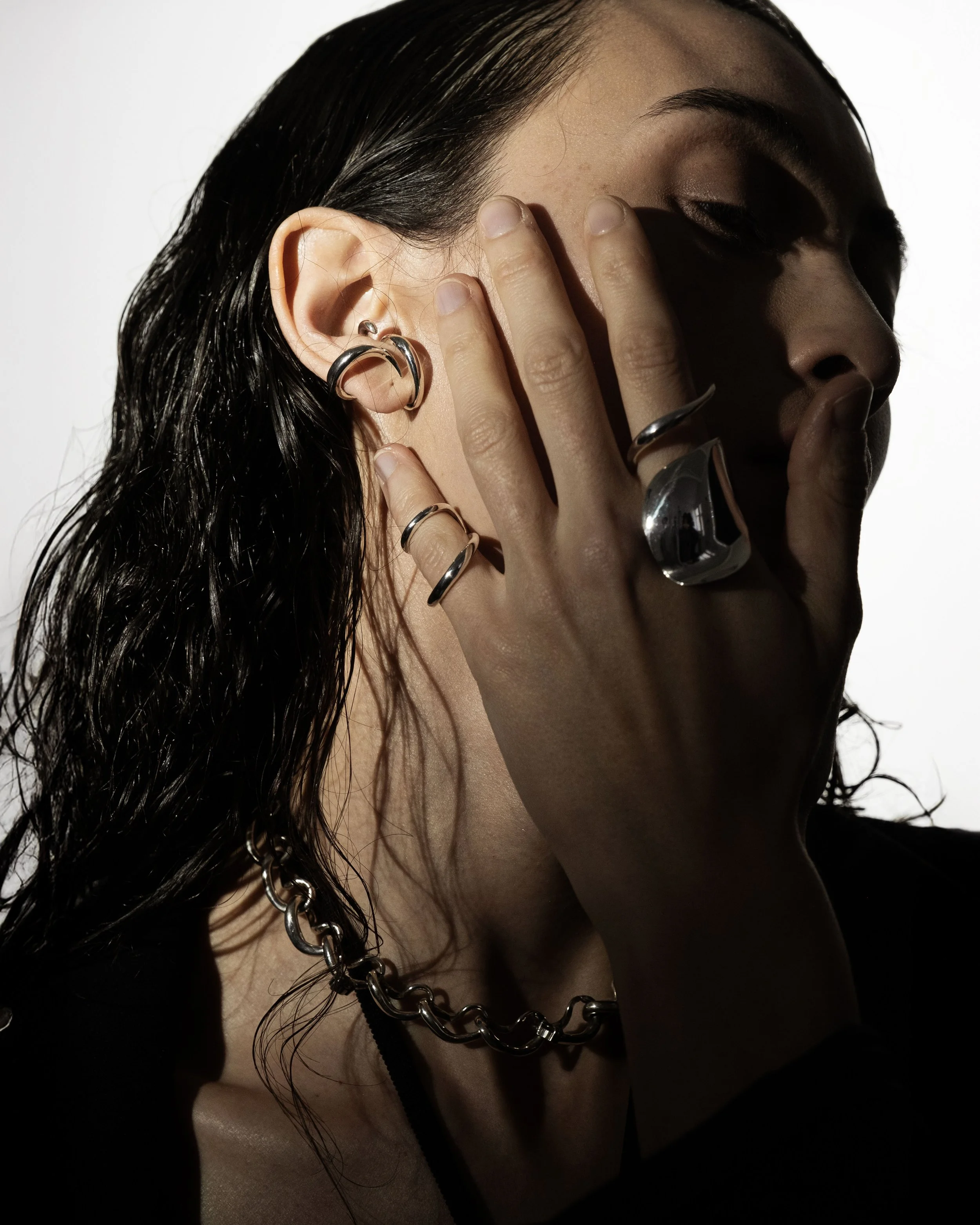 A woman with dark, wet hair wearing silver jewelry covering part of her face with her hand.