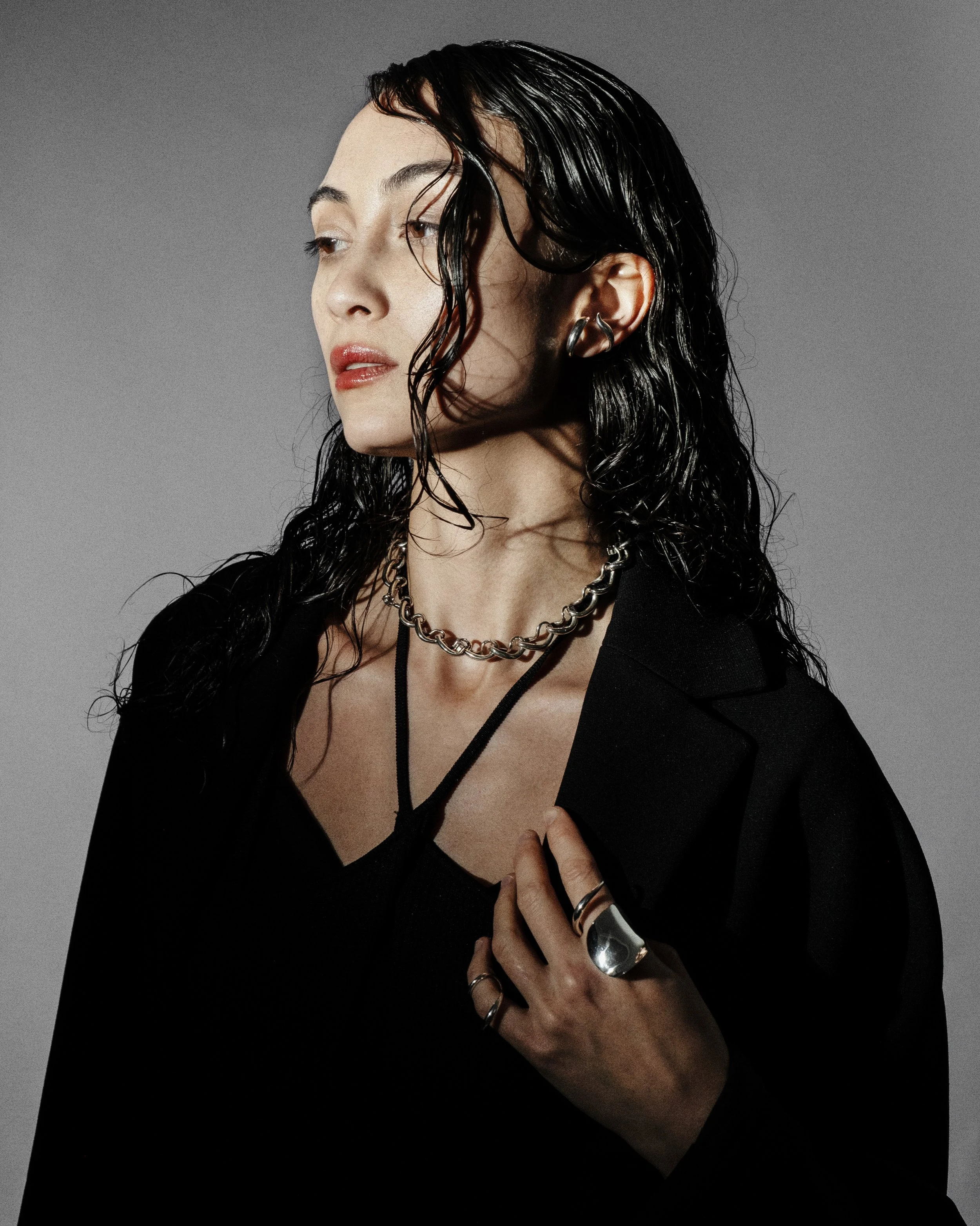 A woman with dark, wavy hair and light skin wearing a black blazer and jewelry, including a thick chain necklace, rings, and earrings, posing against a gray background.
