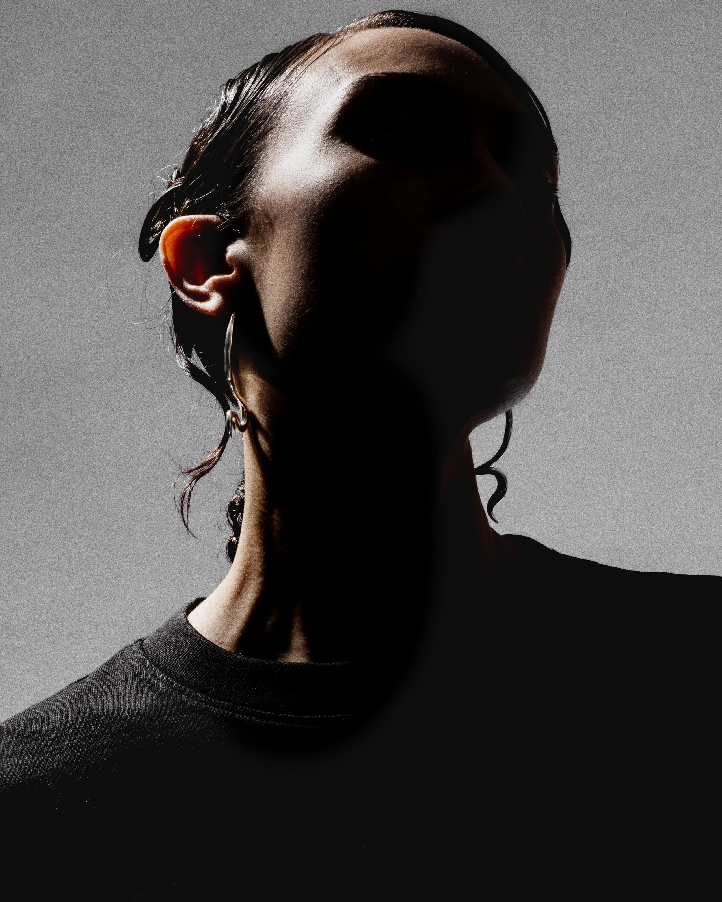 A woman with wet hair, wearing earrings, wearing a dark shirt, standing against a gray background, with dramatic lighting creating shadows on her face.