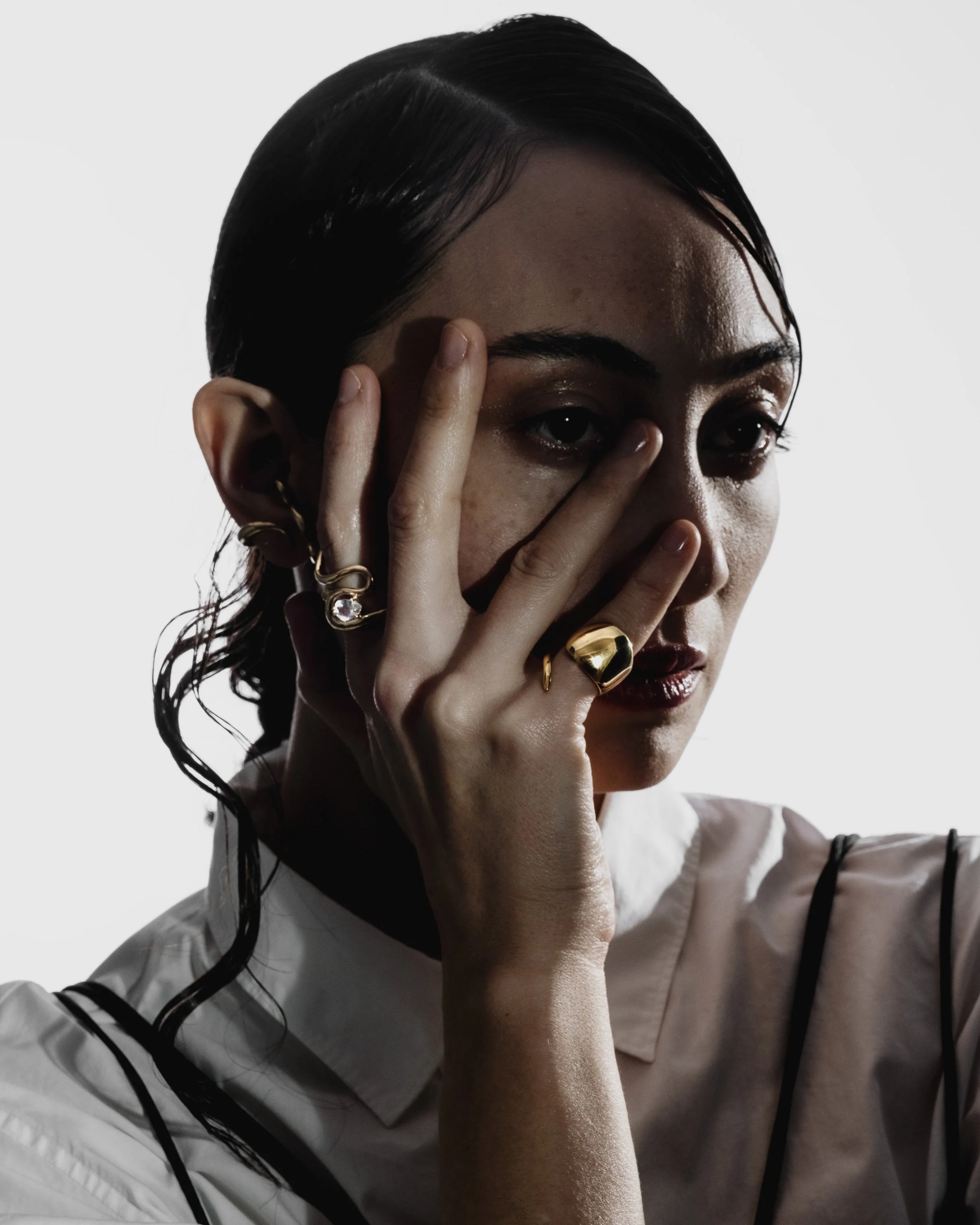 A woman with dark wet hair, wearing jewelry, touching her face with her fingers, and dressed in a white shirt with black suspenders against a white background.