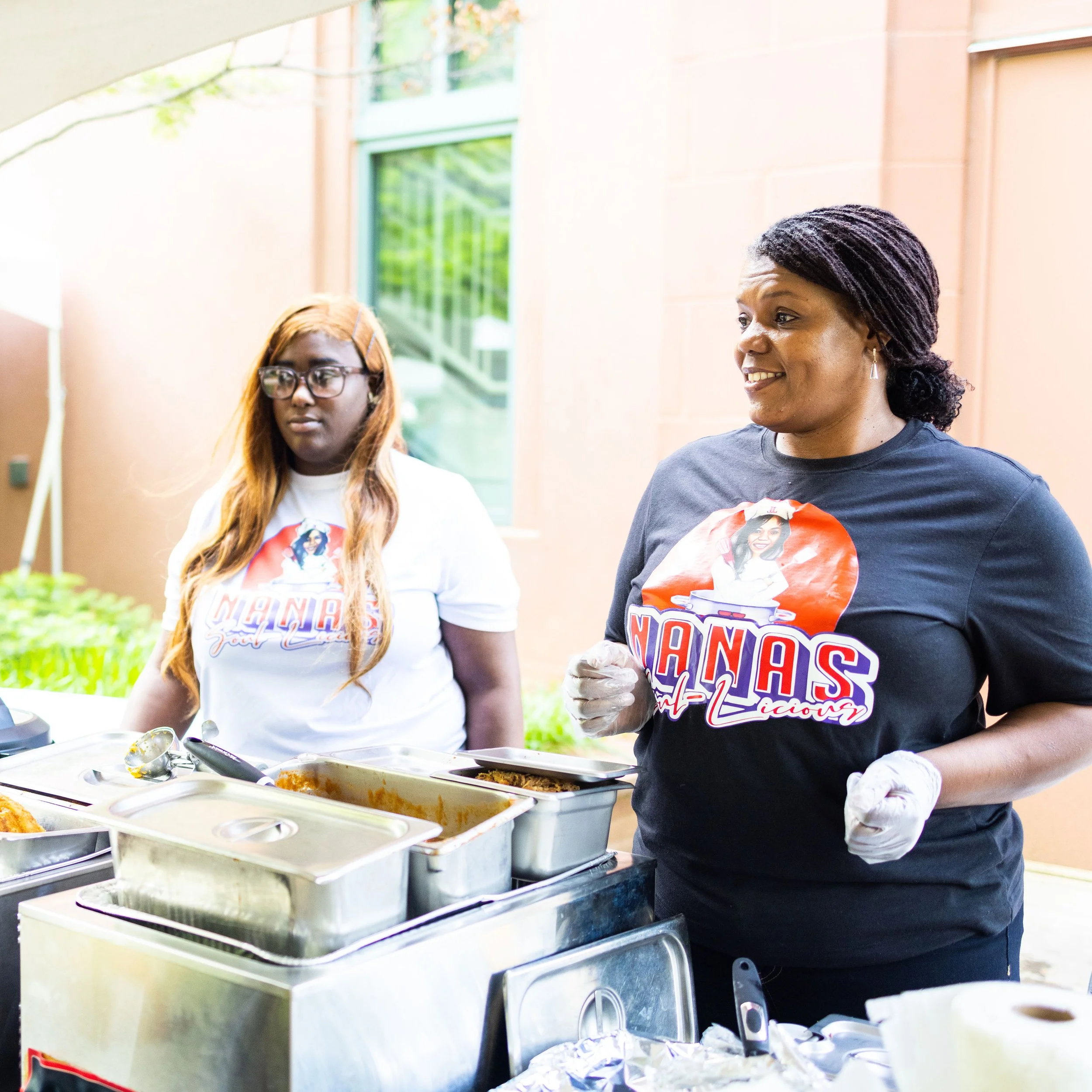 Two women standing behind a food serving station, serving food at an outdoor event, with one woman smiling and wearing a black shirt, and the other woman with glasses and long hair, wearing a white shirt.