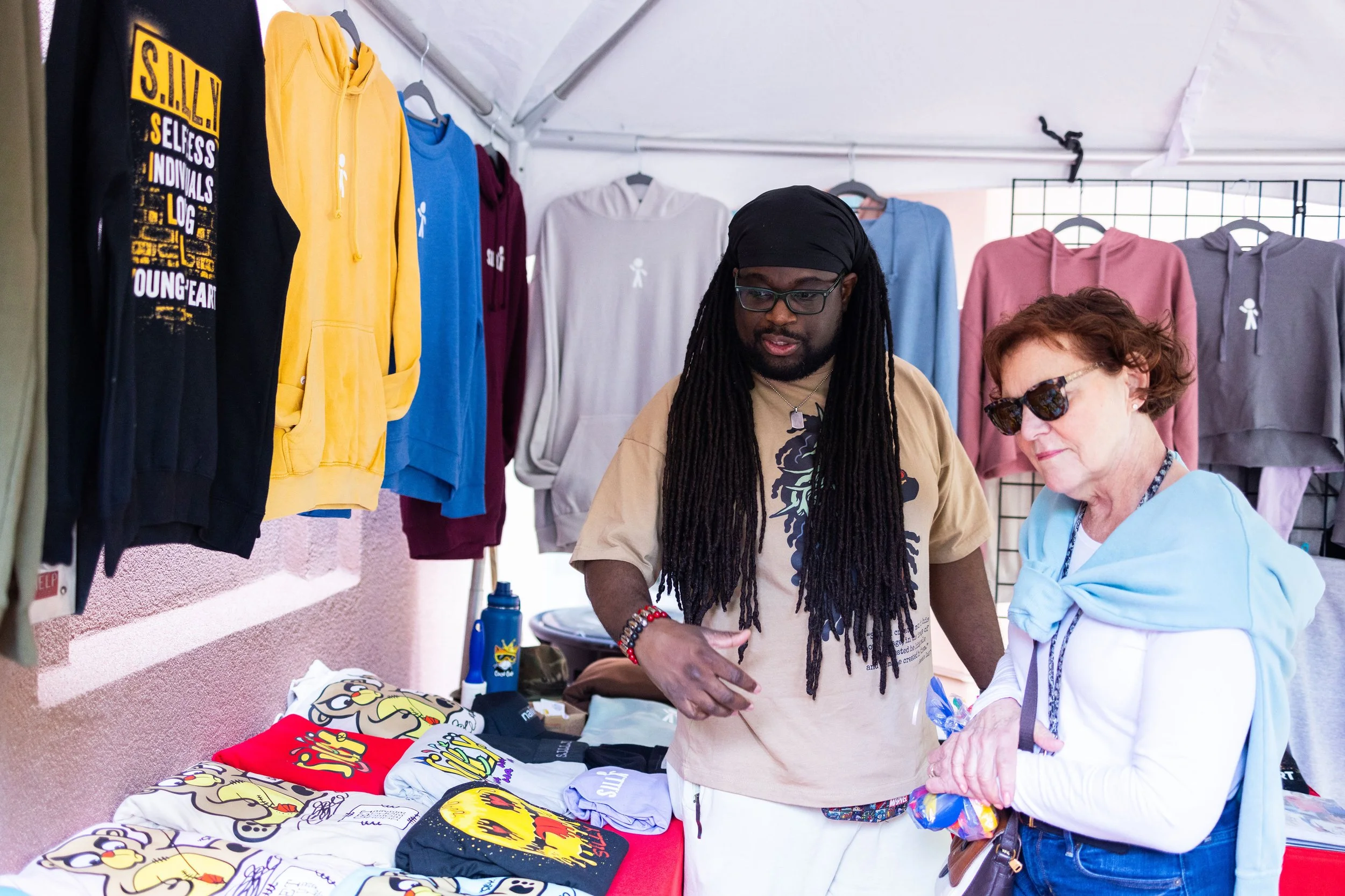 Two women shopping at a booth with colorful hoodies and t-shirts, some with cartoon or comic designs, inside a tent.