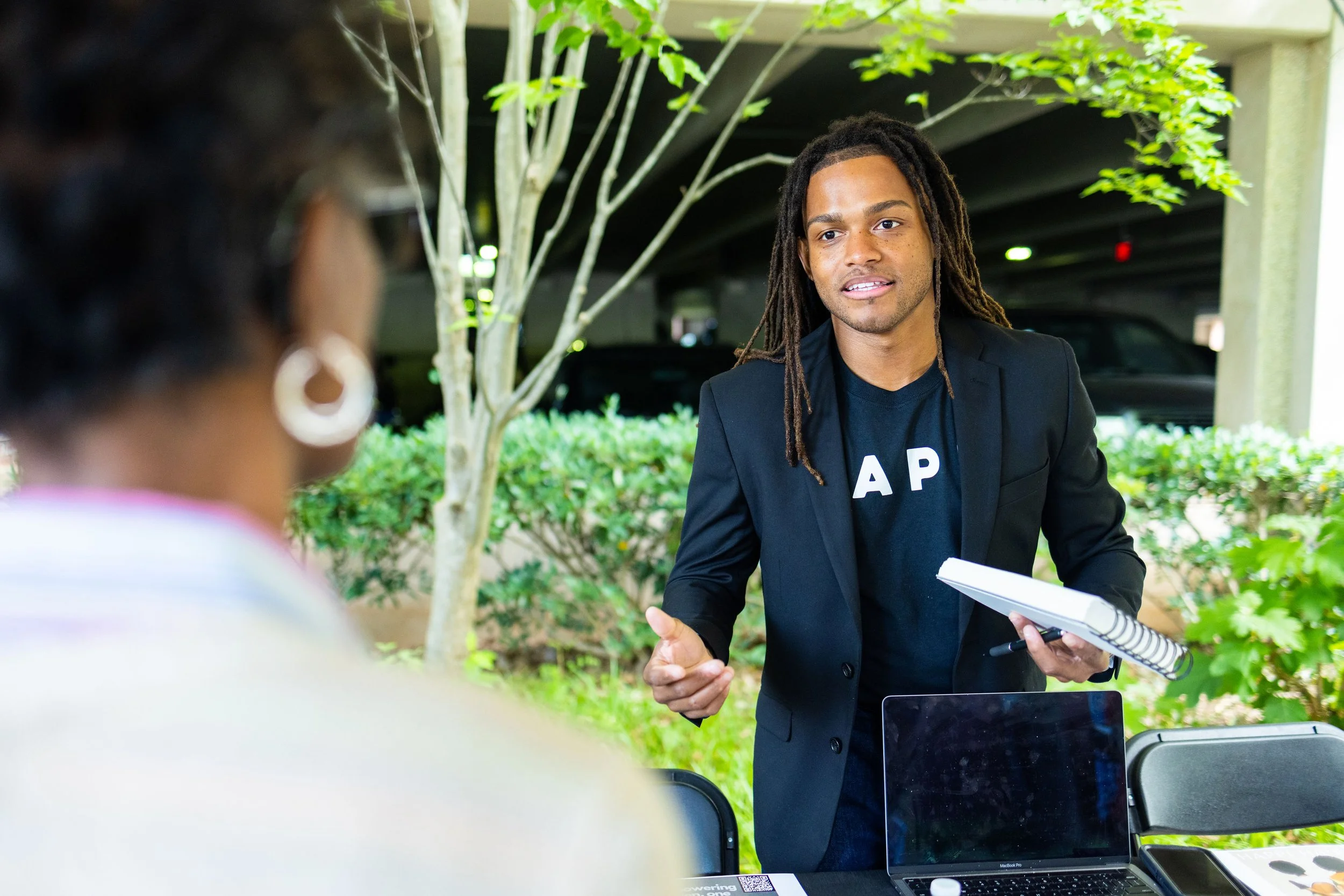 A young man with long dreadlocks, wearing a black blazer and a black T-shirt with white letters, is talking to a woman at an outdoor event table. The table has a laptop, notebooks, and other items, with greenery and parking structure in the backgroun