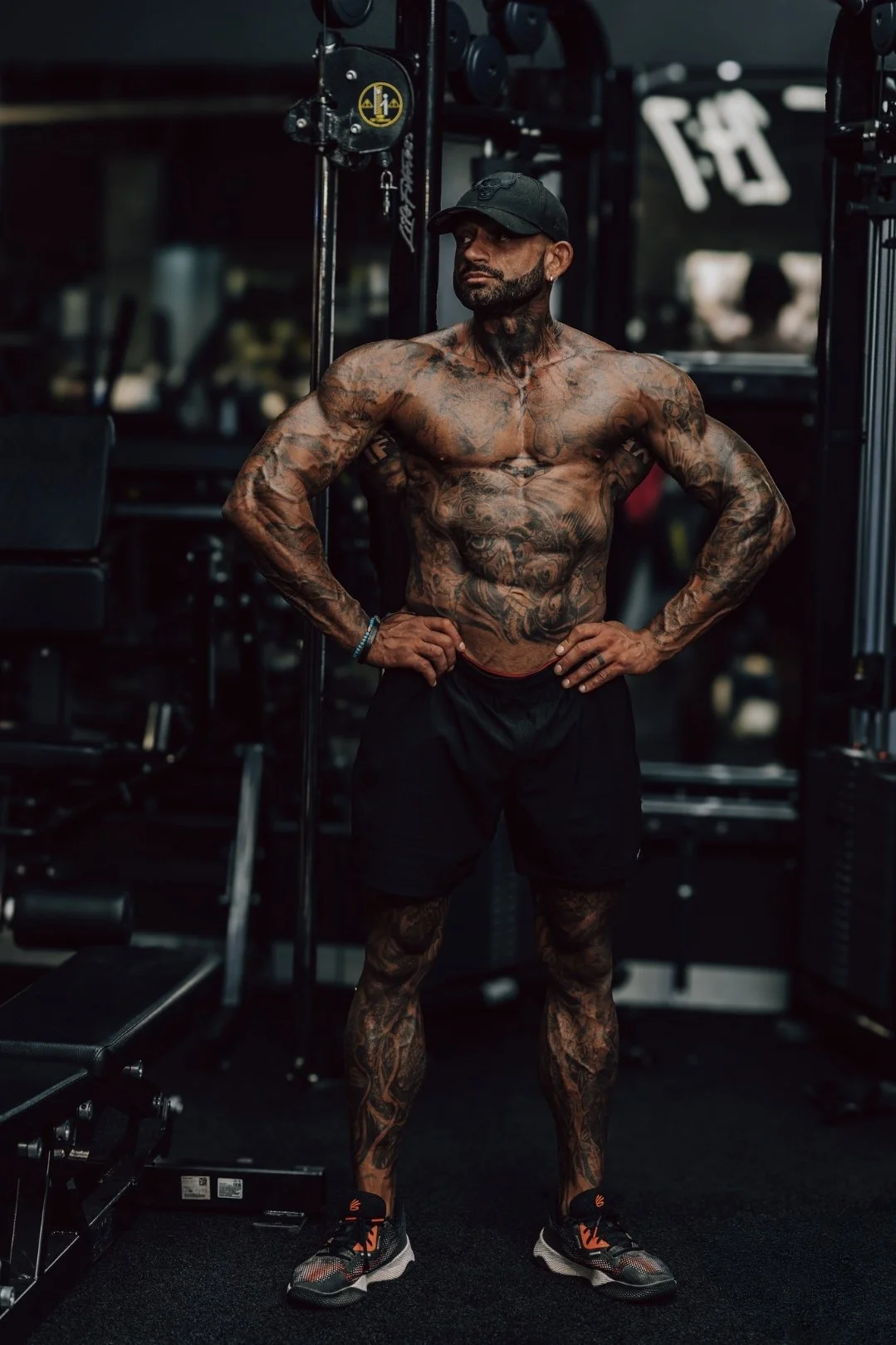 Tattooed man wearing a black cap, shorts, and sneakers standing with hands on hips in a gym.