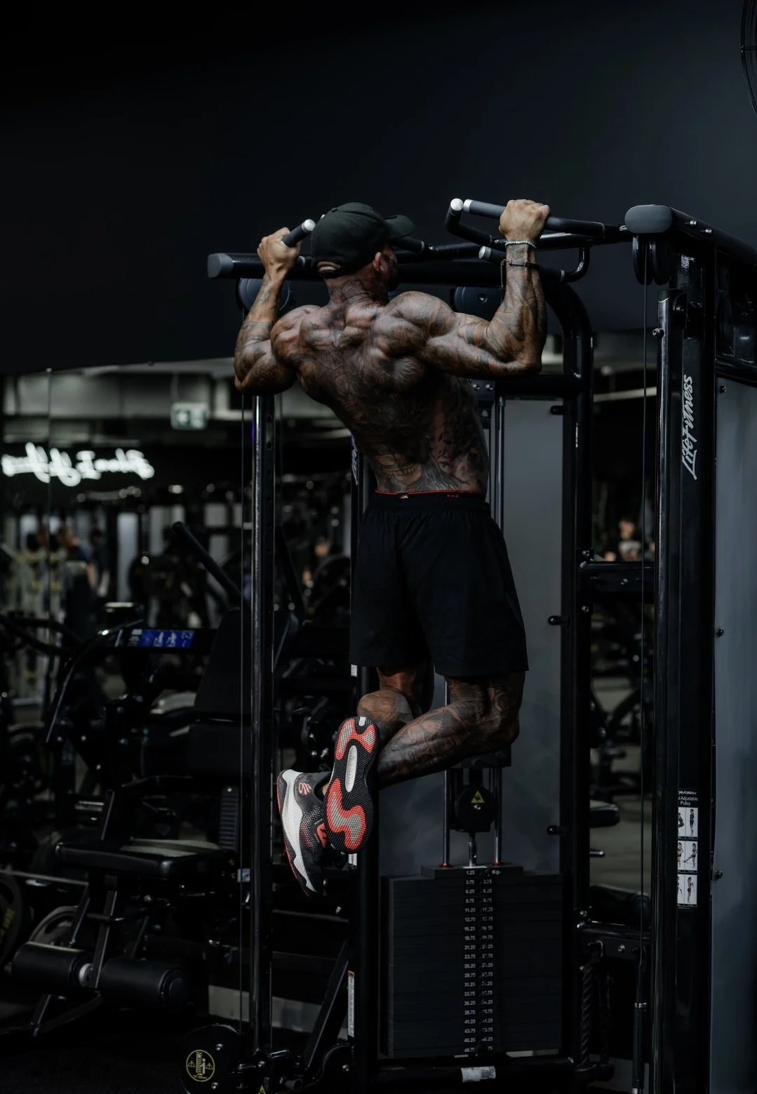 A muscular man with tattoos is doing pull-ups on a fitness rack in a gym, wearing black shorts, a black cap, and sneakers.