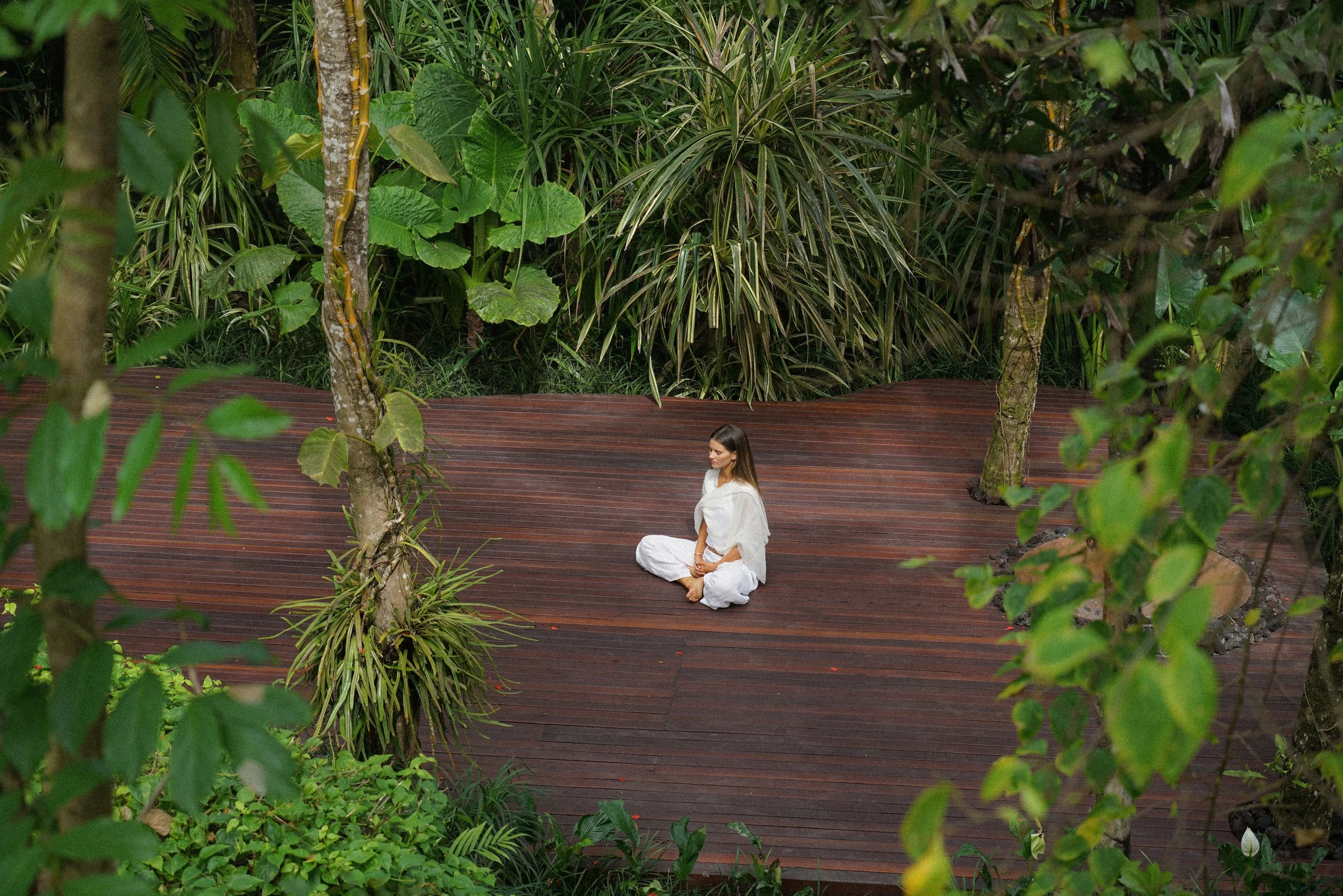 A woman in white clothing sitting cross-legged on a wooden deck surrounded by lush green tropical plants.