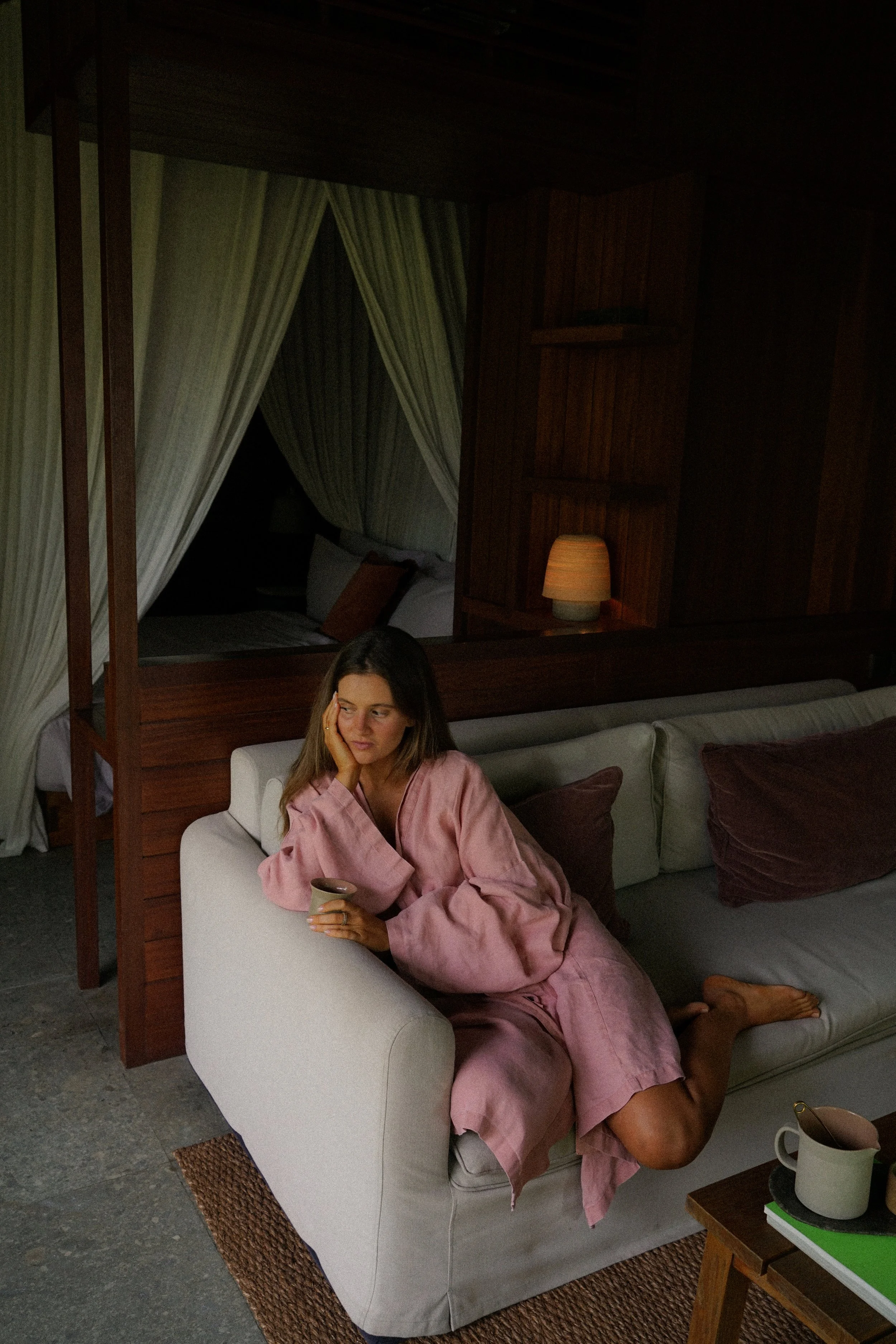 A woman in pink pajamas sits on a white sofa holding a cup, with a thoughtful expression. Behind her is a wooden partition with a lamp, and a bed with curtains is visible in the background.
