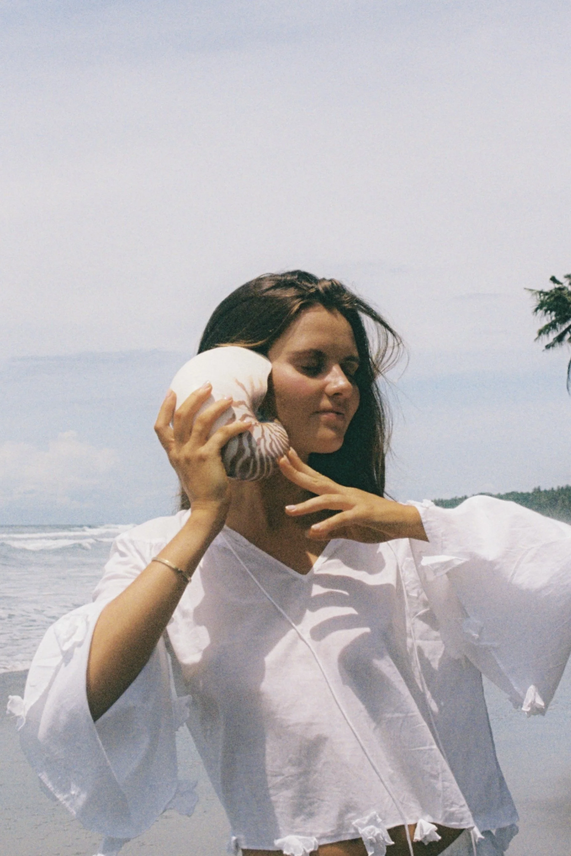 A woman holding a seashell up to her ear on the beach with the ocean and sky in the background.