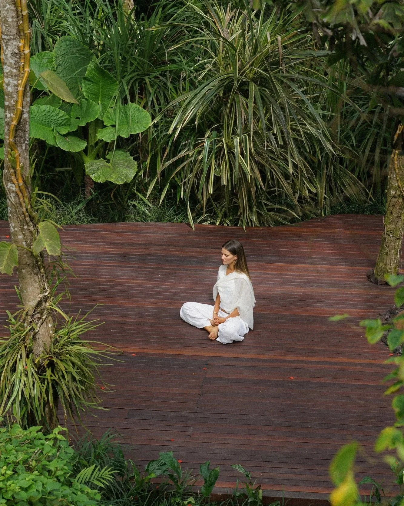 A woman sitting cross-legged on a wooden platform in a lush, green tropical garden, meditating.