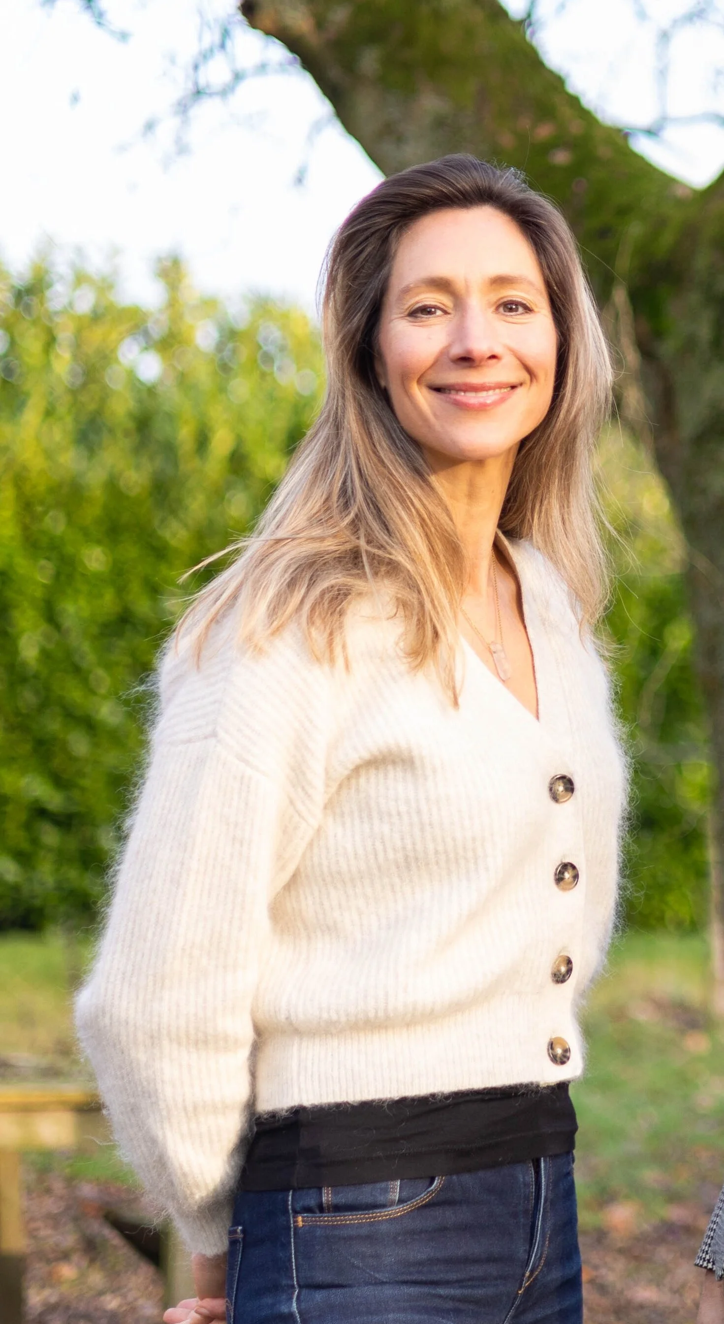 Close-up van een glimlachende vrouw met bruin haar, gekleed in een cremekleurige cardigan met knopen en een zwarte top eronder, staand buiten voor een boom en groene achtergrond.
