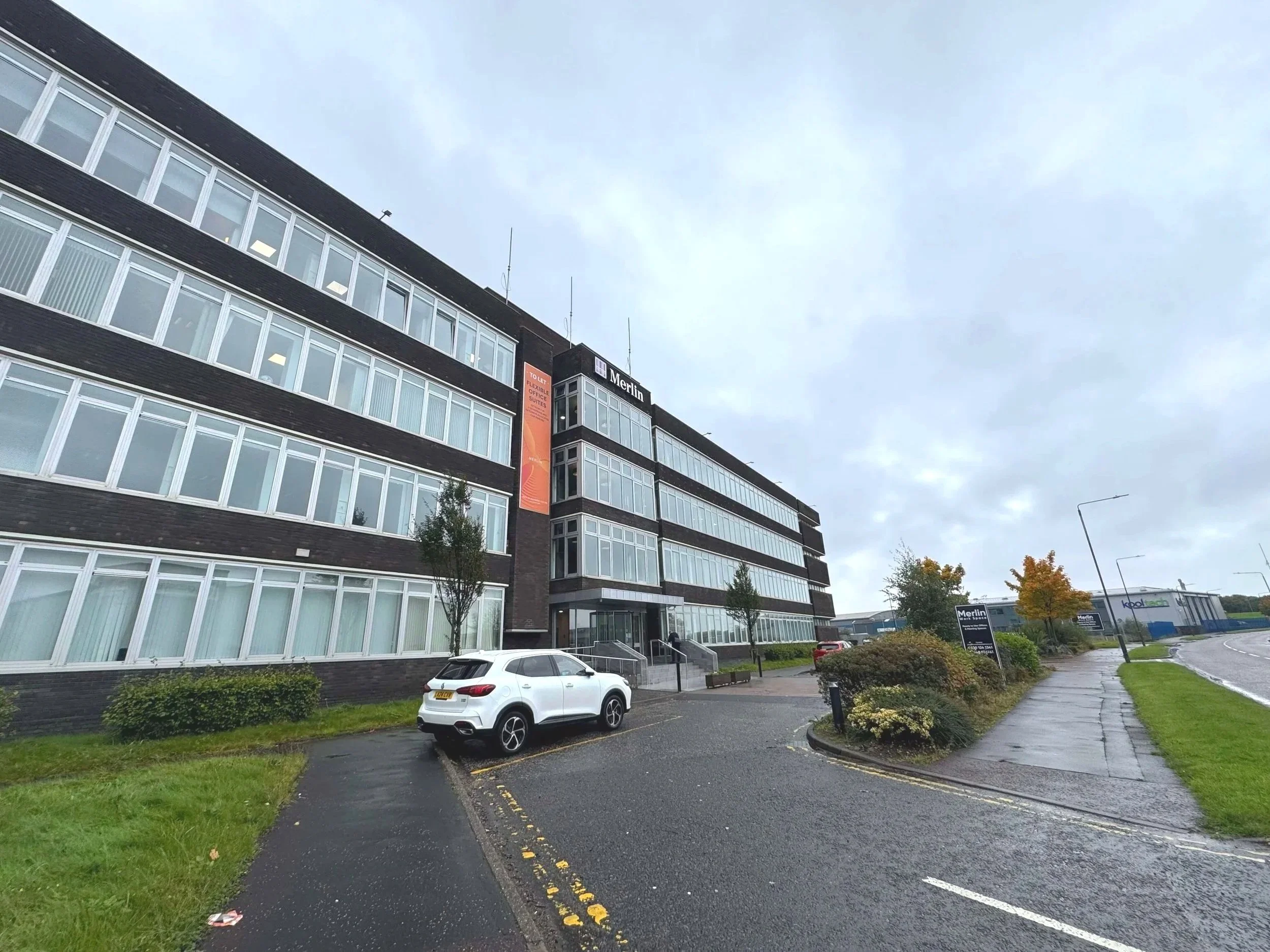 A modern multi-story office building with a dark brick facade and large windows, with a parking lot and some trees in front. The sky is overcast and the ground appears wet.
