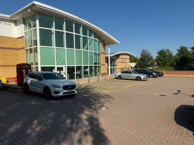 Empty parking lot in front of a modern building with large glass windows, several parked cars, and trees in the background on a sunny day.