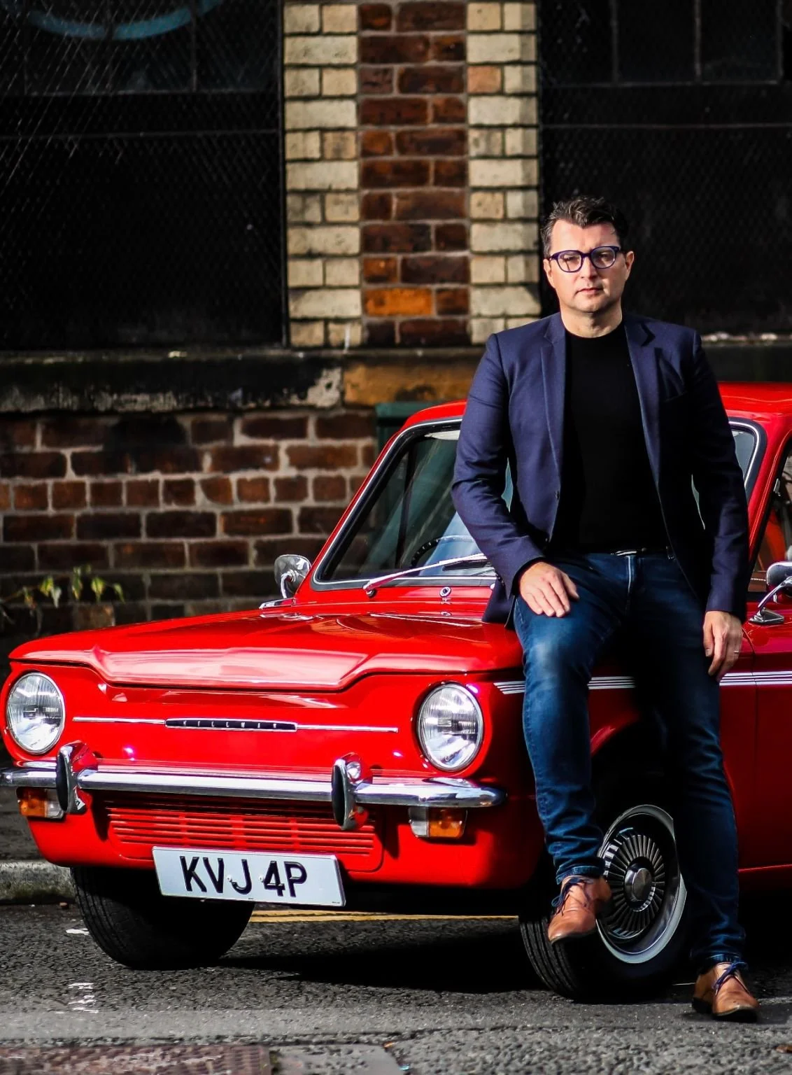 A man wearing glasses, a black shirt, and a navy blazer leaning against a red vintage car parked on a street in front of a brick wall.