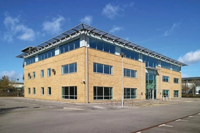 A modern, multi-story commercial building with brick and glass facade, large windows, and a metal awning on top, situated in a parking lot under a blue sky with clouds.