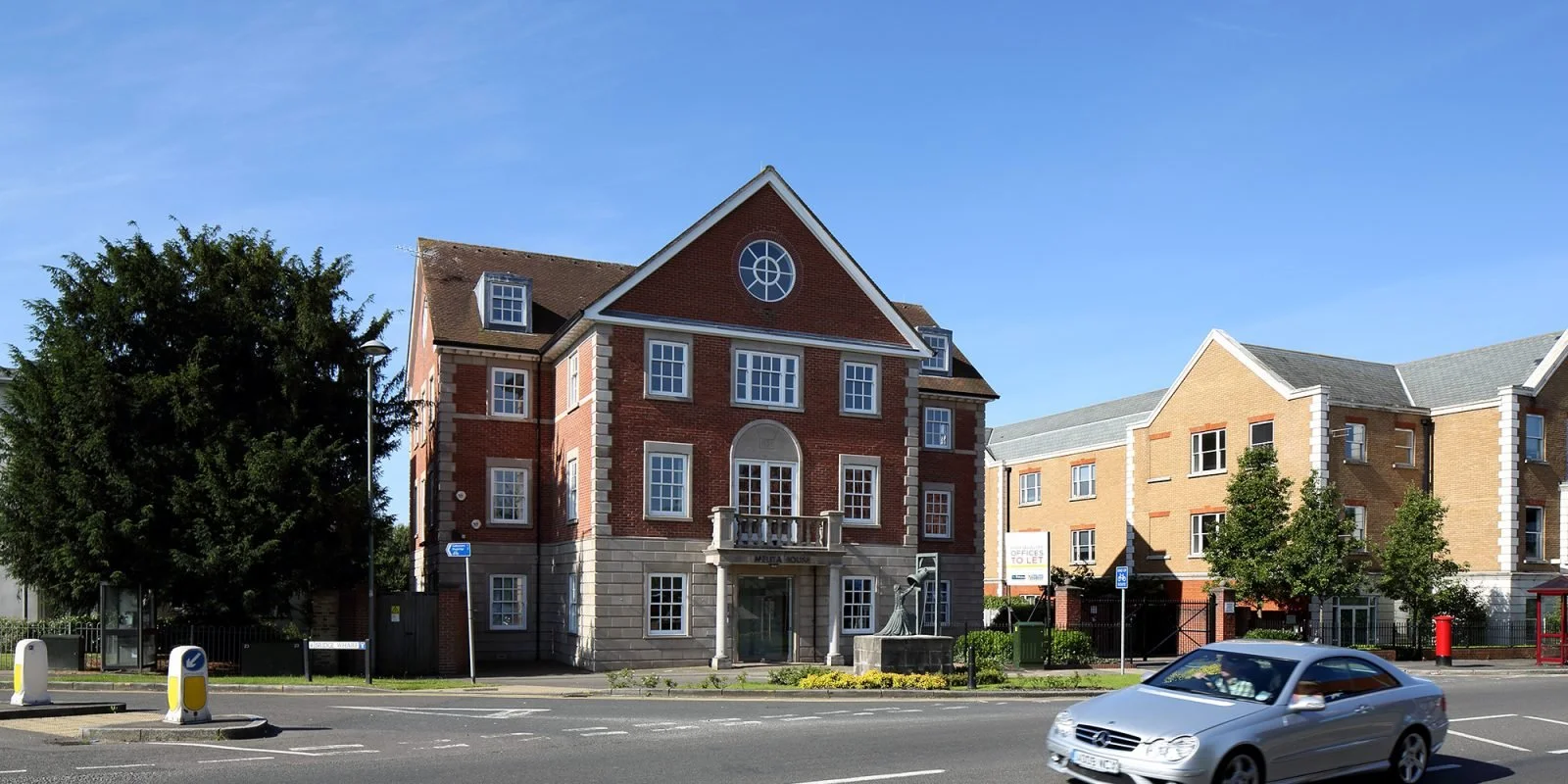 Street view with a large brick residential building, trees, parked car, and a clear blue sky.