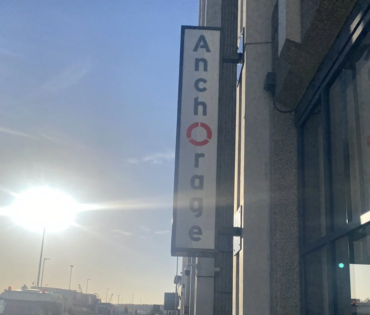 Sign reading 'Anchorage' with a red circle symbol, on a building's exterior wall, with parked vehicles and a bright sun in the clear sky.