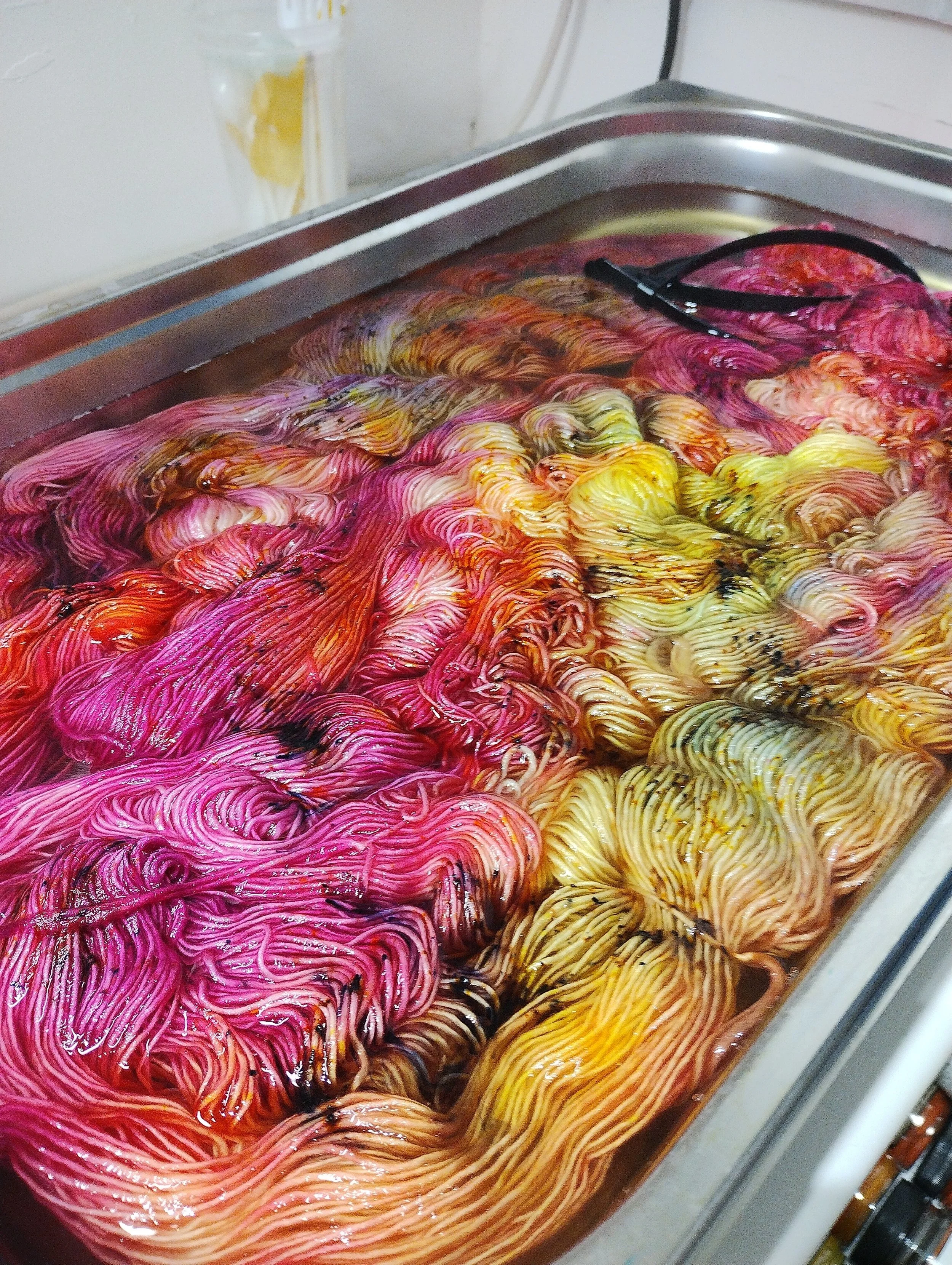 Colorful dyed yarns in shades of pink, yellow, and orange submerged in water on a metal tray.