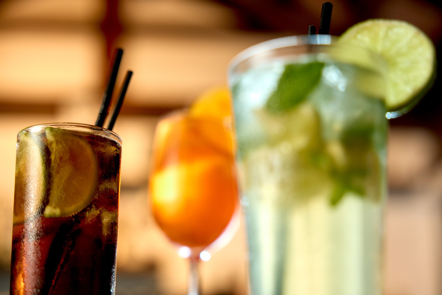Three colorful cocktails with lime, orange, and mixed fruit garnishes, shot in close-up with a blurred background.