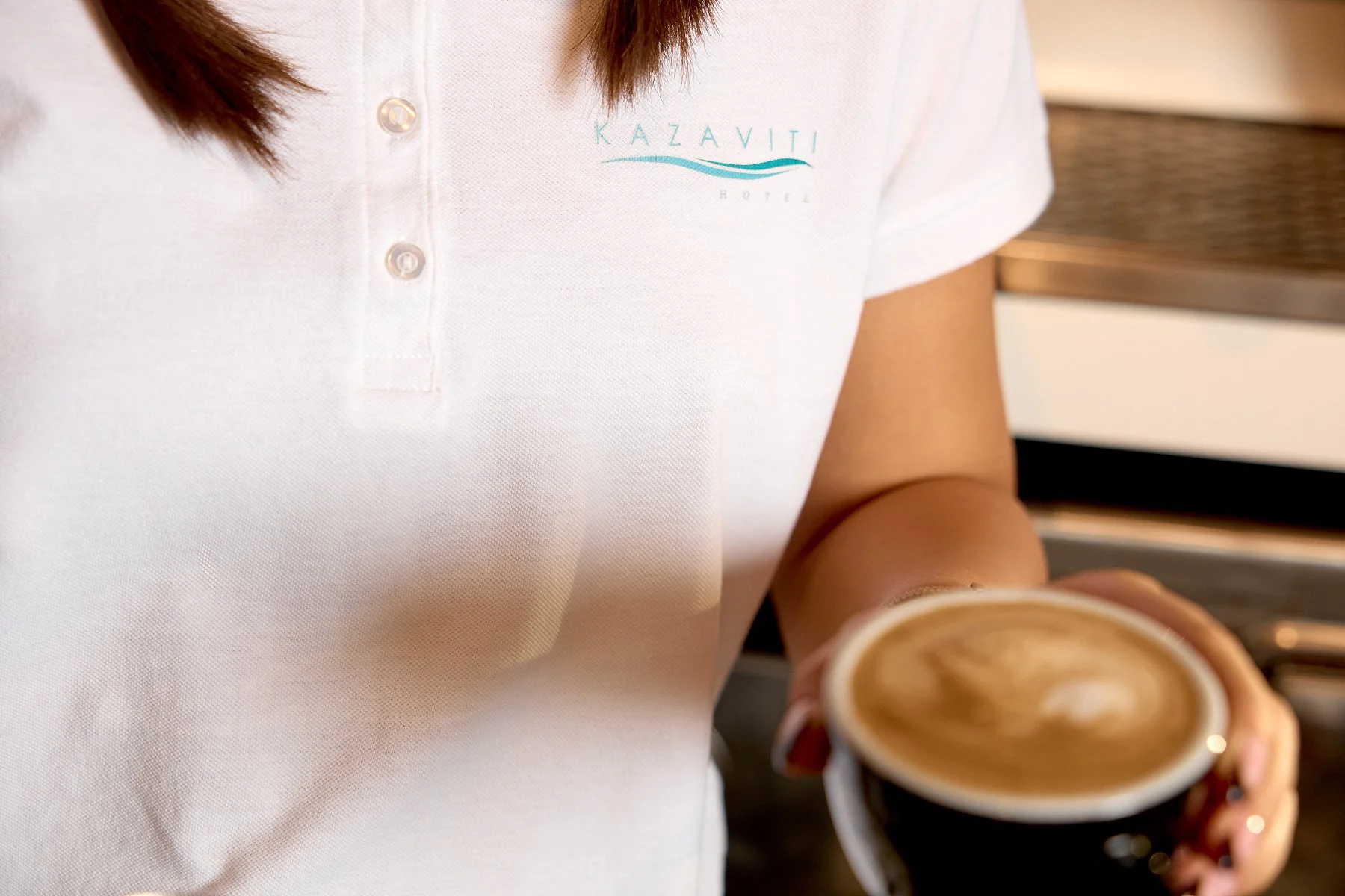 A person wearing a Kazaviti Hotel polo shirt holding a cup of coffee with latte art, standing in a coffee shop.