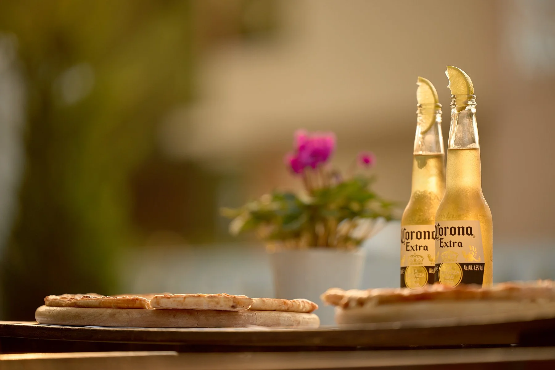 Two bottles of Corona Extra beer with lime wedges on top, placed on a wooden surface with a blurred background including a potted plant with purple flowers.