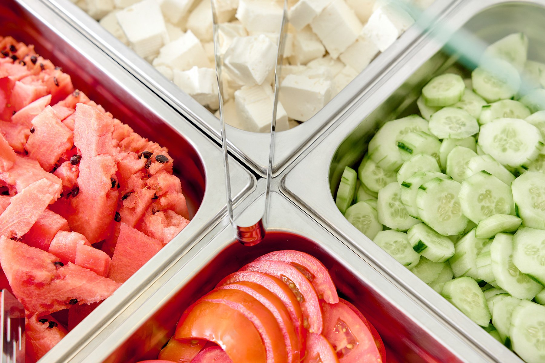 A close-up of a salad bar with sections containing chopped watermelon, diced tofu, sliced cucumbers, and sliced tomatoes.