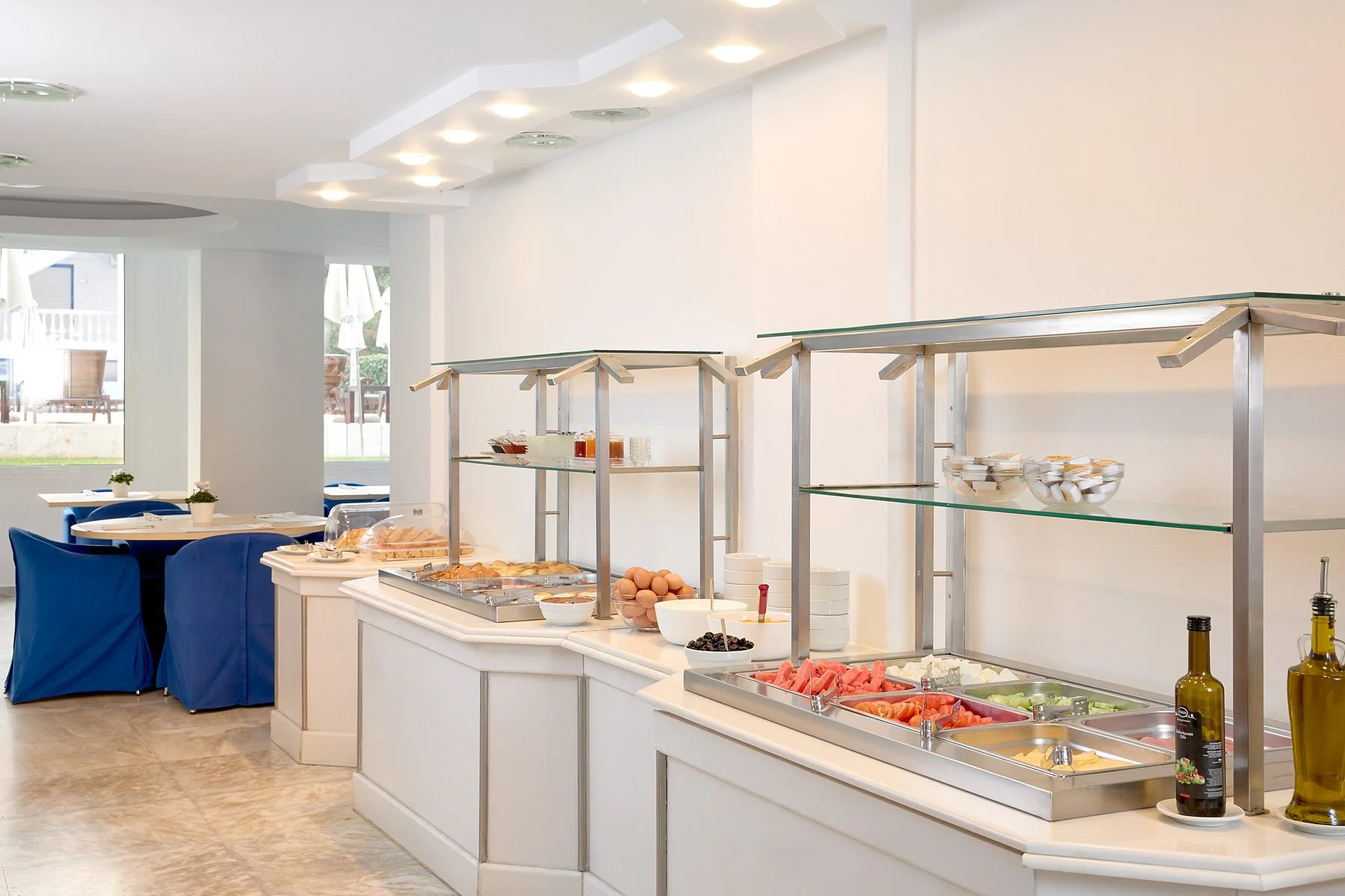 Buffet area with an assortment of breakfast foods including cereal, fruit, eggs, and pastries in a bright, clean dining area with blue-covered chairs and white tables.
