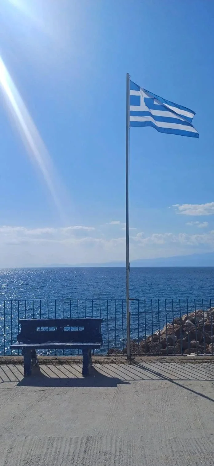 Blue and white Greek flag on a flagpole beside the ocean, with a bench and rocks on a sidewalk.