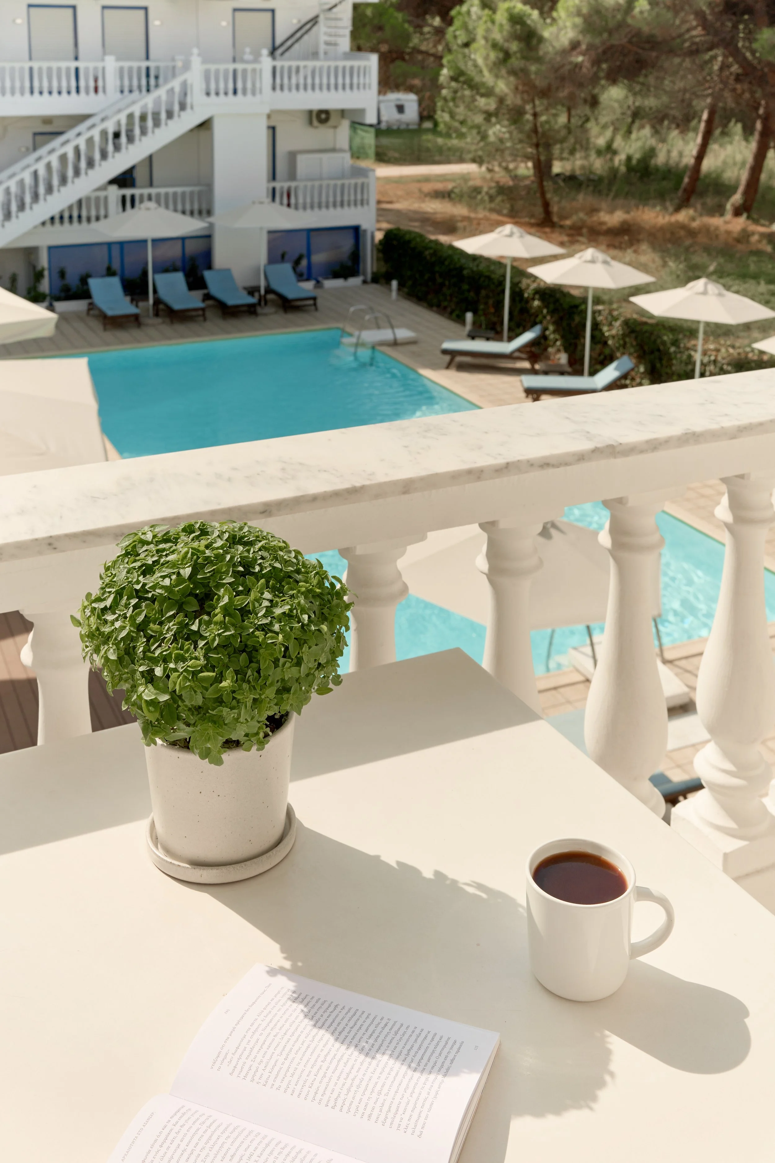 A white table on a balcony with a potted green plant, a cup of coffee, and an open book. In the background, there is a swimming pool with lounge chairs and umbrellas, surrounded by trees and greenery.