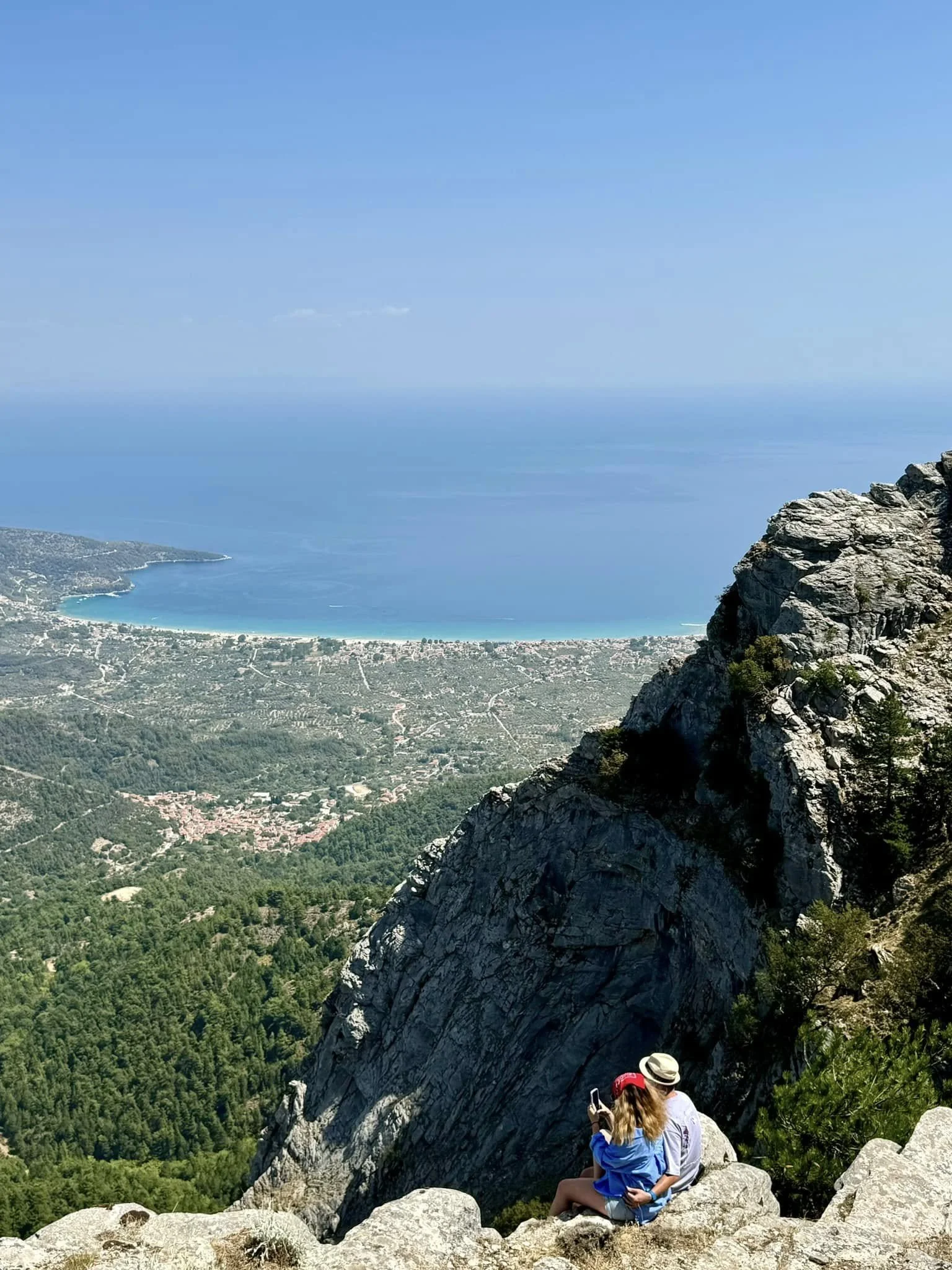 A couple sitting on rocks on a mountain ledge overlooking a coastal landscape with a bay, forest, and ocean in the background.