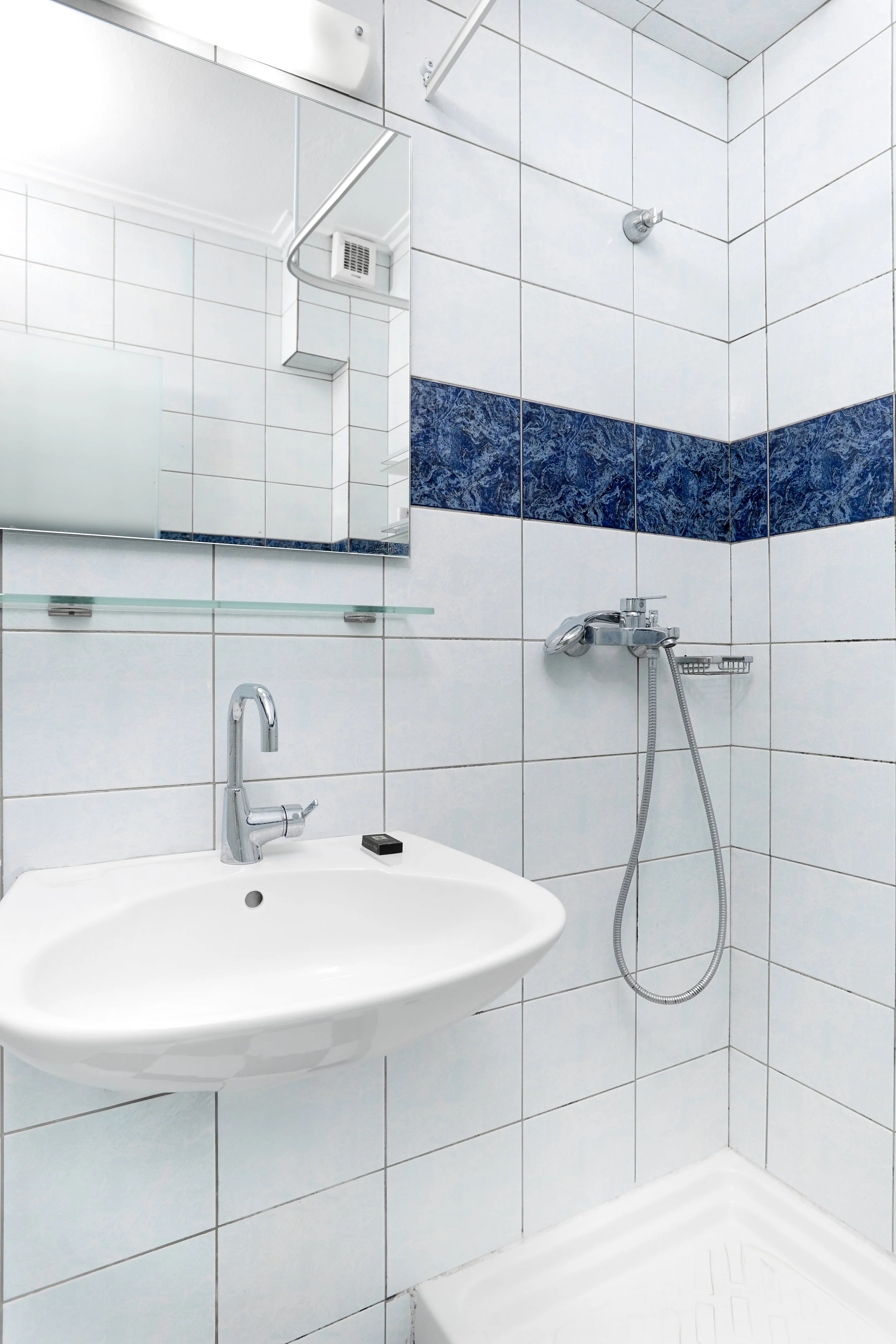 View of a bathroom with white tiled walls, a small white ceramic sink with a chrome faucet, a rectangular mirror, a small soap dish, and a shower area with a wall-mounted showerhead and a flexible hose.