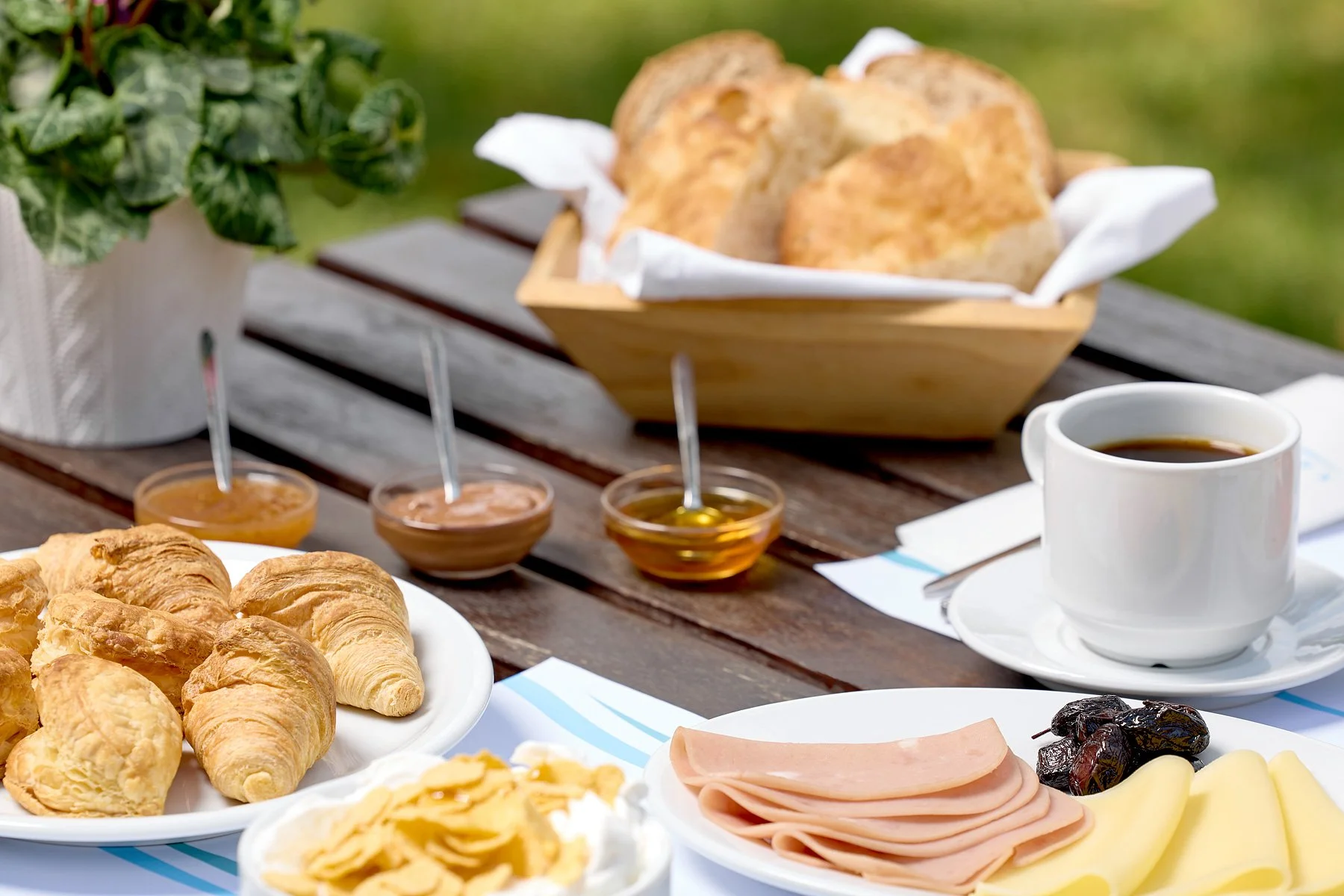 Breakfast spread on a wooden table including croissants, bread, honey, jam, honey, a cup of coffee, slices of ham, cheese, and dates, with a potted plant in the background.