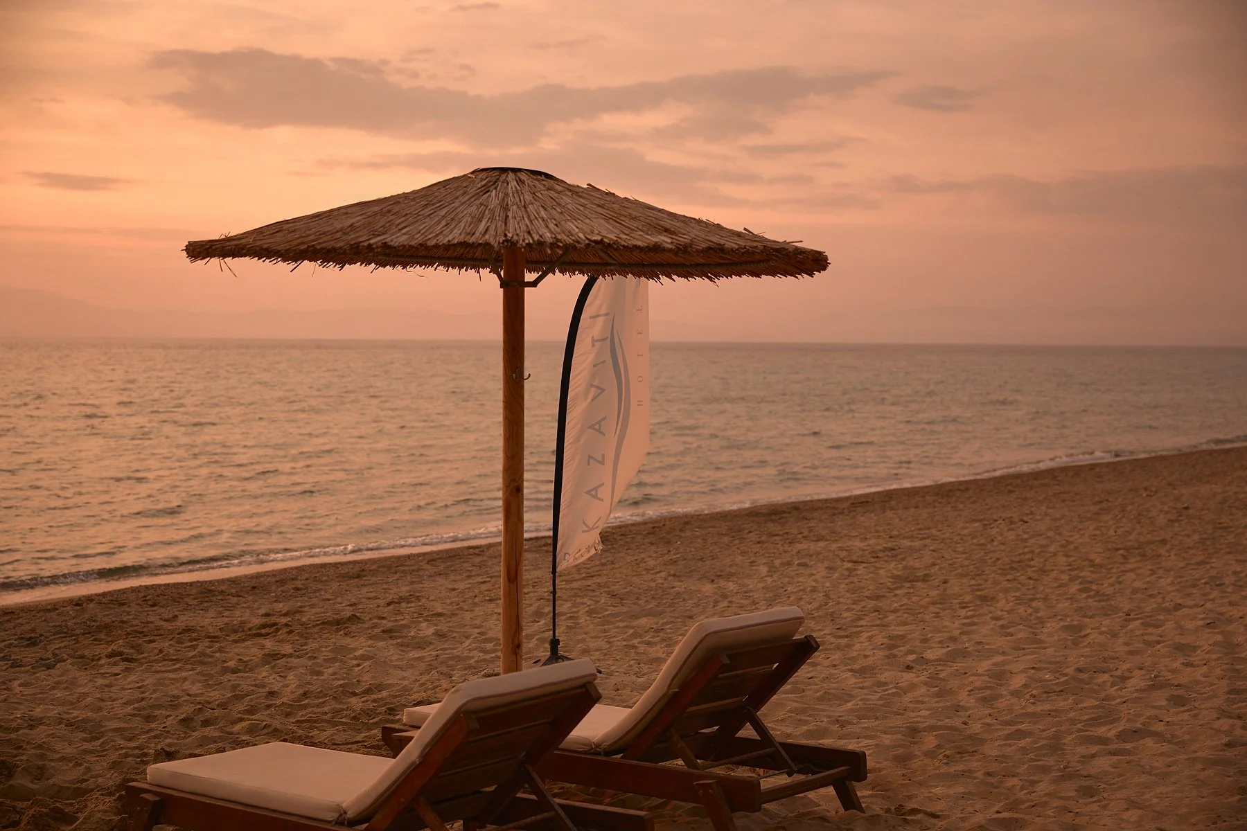 Empty beach with two lounge chairs under a thatched umbrella and a flag, overlooking the ocean at sunset.