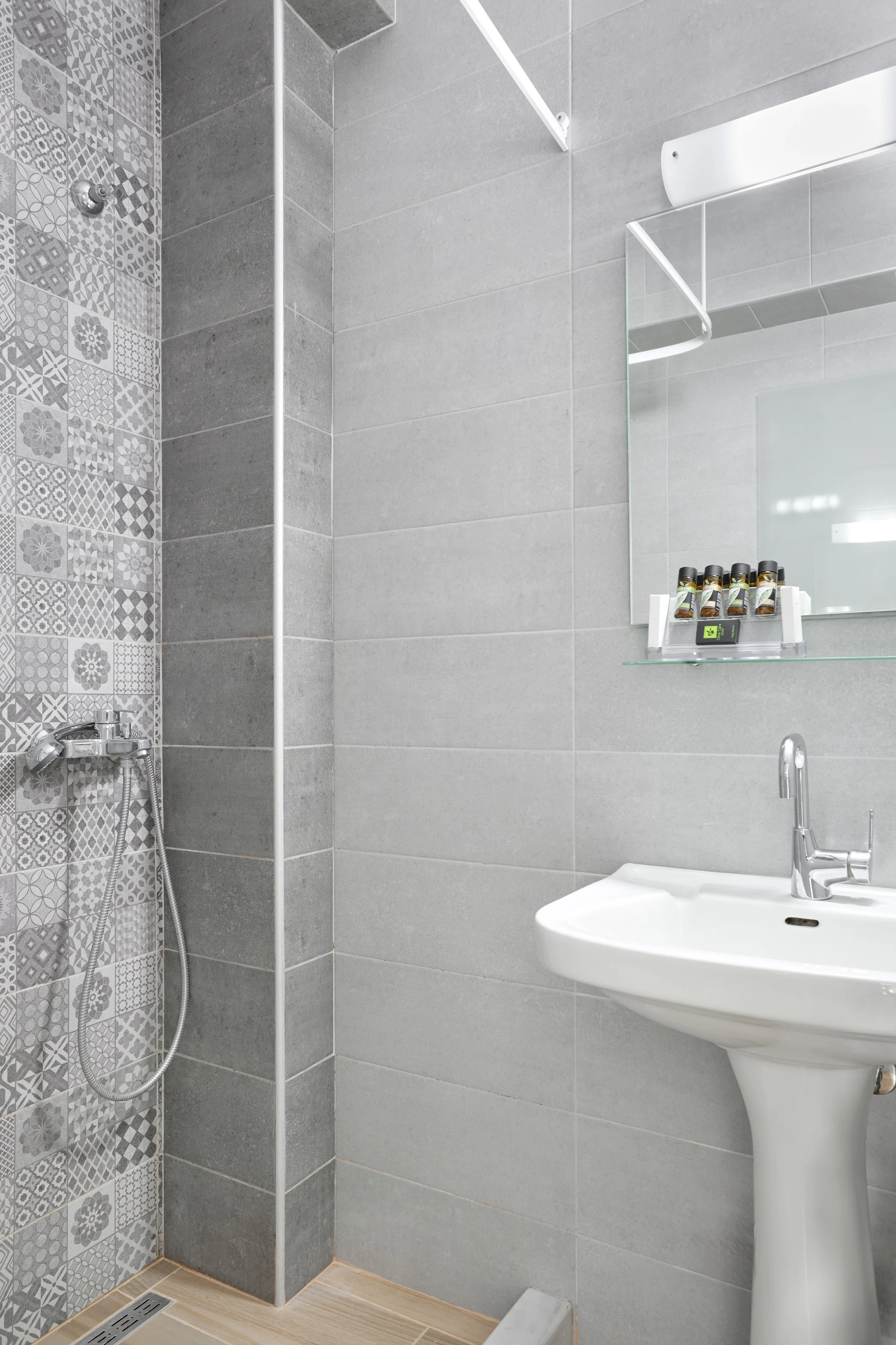 A modern small bathroom with a shower area on the left, featuring patterned grey tiles, a white pedestal sink on the right, a large rectangular mirror above the sink, and a glass shelf with multiple small bottles. The walls are tiled in grey, and the