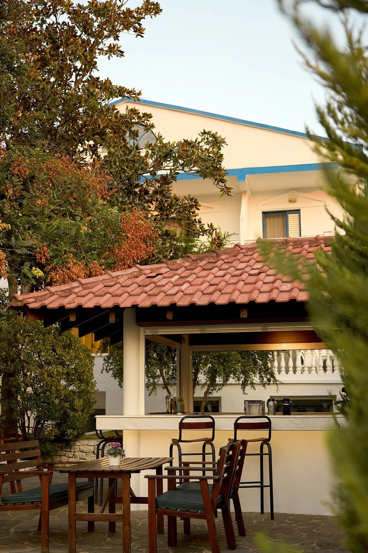 An outdoor patio area featuring a bar with a tiled roof, wooden chairs, a small table with a flowerpot, and surrounding greenery. In the background, there is a white building with blue accents and a balcony.