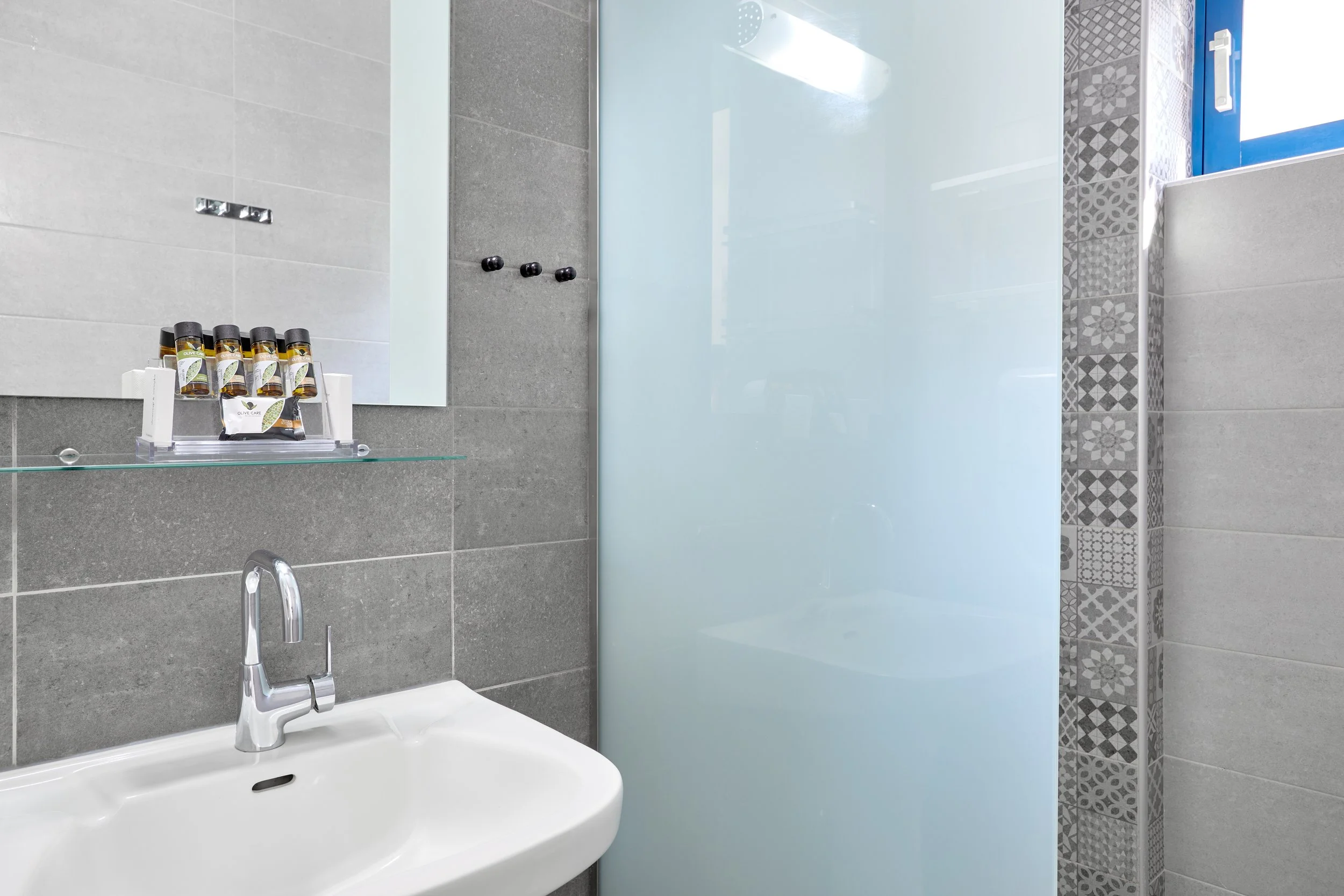 Modern bathroom with gray tiled walls, a white sink with a chrome faucet, a glass shelf holding small bottles, a mirror, a frosted glass shower door, and patterned tiles in the shower area, with a window letting in natural light.