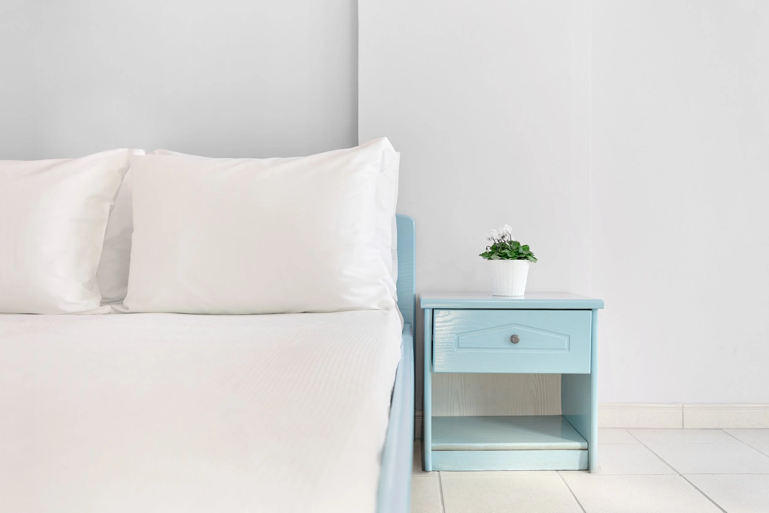 A bedroom with white bedding and pillows, a light blue nightstand with a small drawer and open shelf, and a potted white flowering plant on top of the nightstand, against white walls and tiled floor.