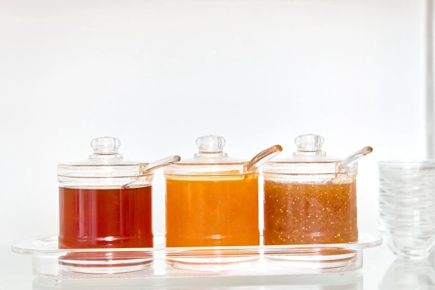 Three glass jars with honey of different colors and a glass of honey sticks on the side, all arranged on a clear tray.