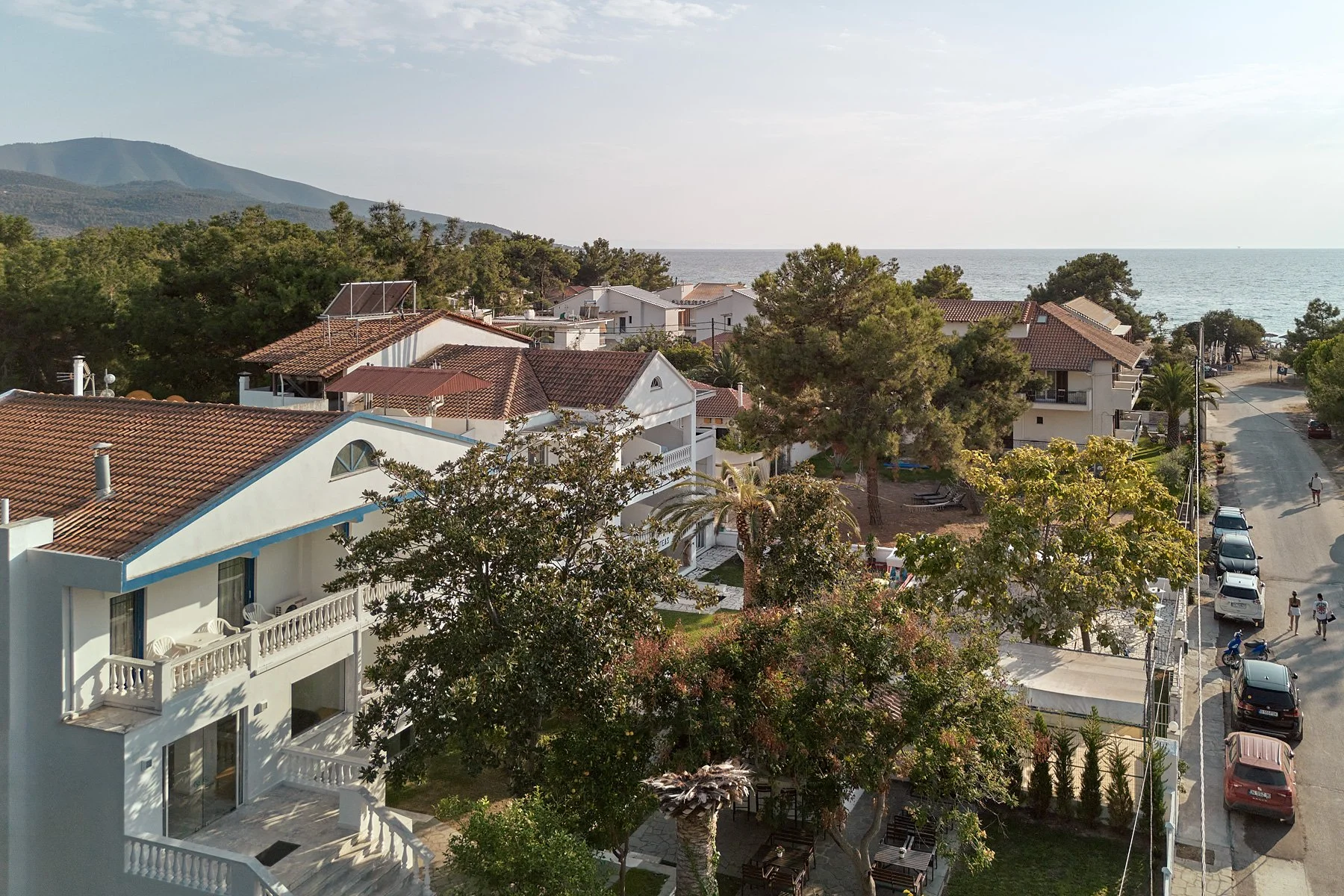 View of a seaside neighborhood with houses, trees, and parked cars, overlooking the ocean and distant mountains.