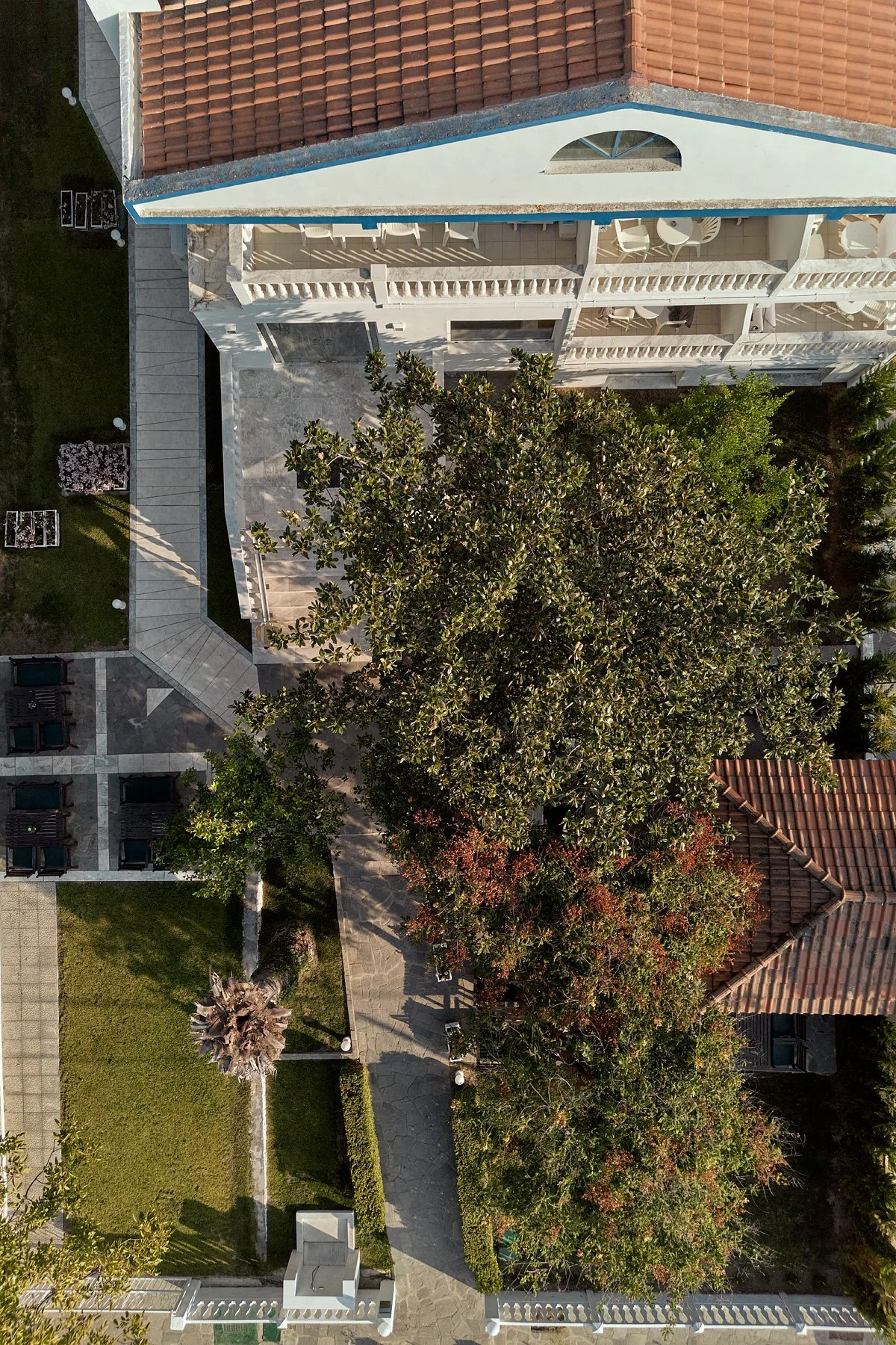 An aerial view of a residential building with a red-tiled roof, balconies with white railings, and surrounding trees, landscaped yard, and paved walkways.