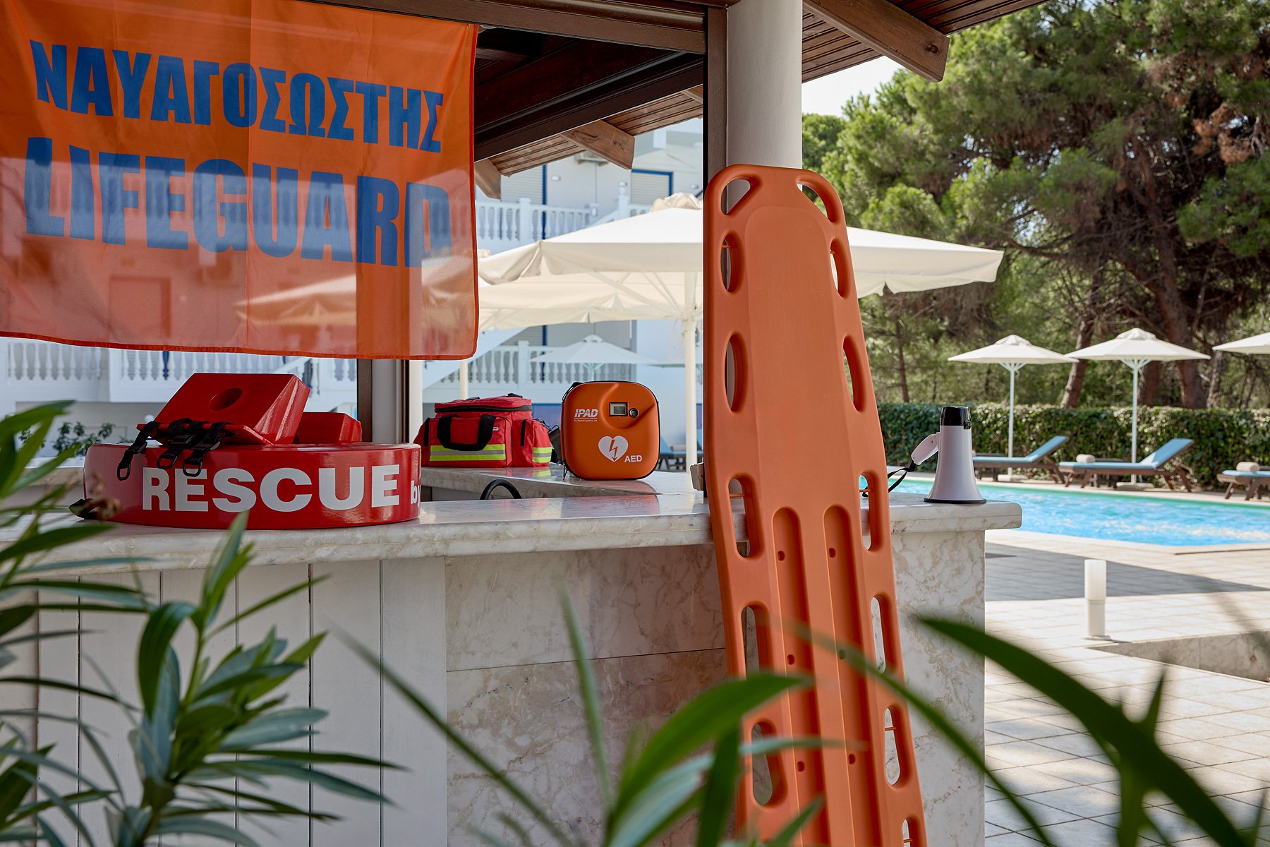 A rescue or lifeguard station at a poolside, featuring rescue gear, a rescue board, an AED device, a first aid kit, and lounge chairs with umbrellas in the background.