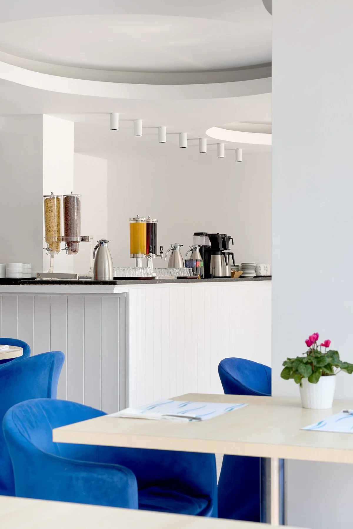 Interior of a dining area with blue chairs, a light-colored table, and a potted pink flower. In the background, a white counter with coffee and cereal dispensers, plates, and a coffee machine is visible, with a minimalist ceiling design.
