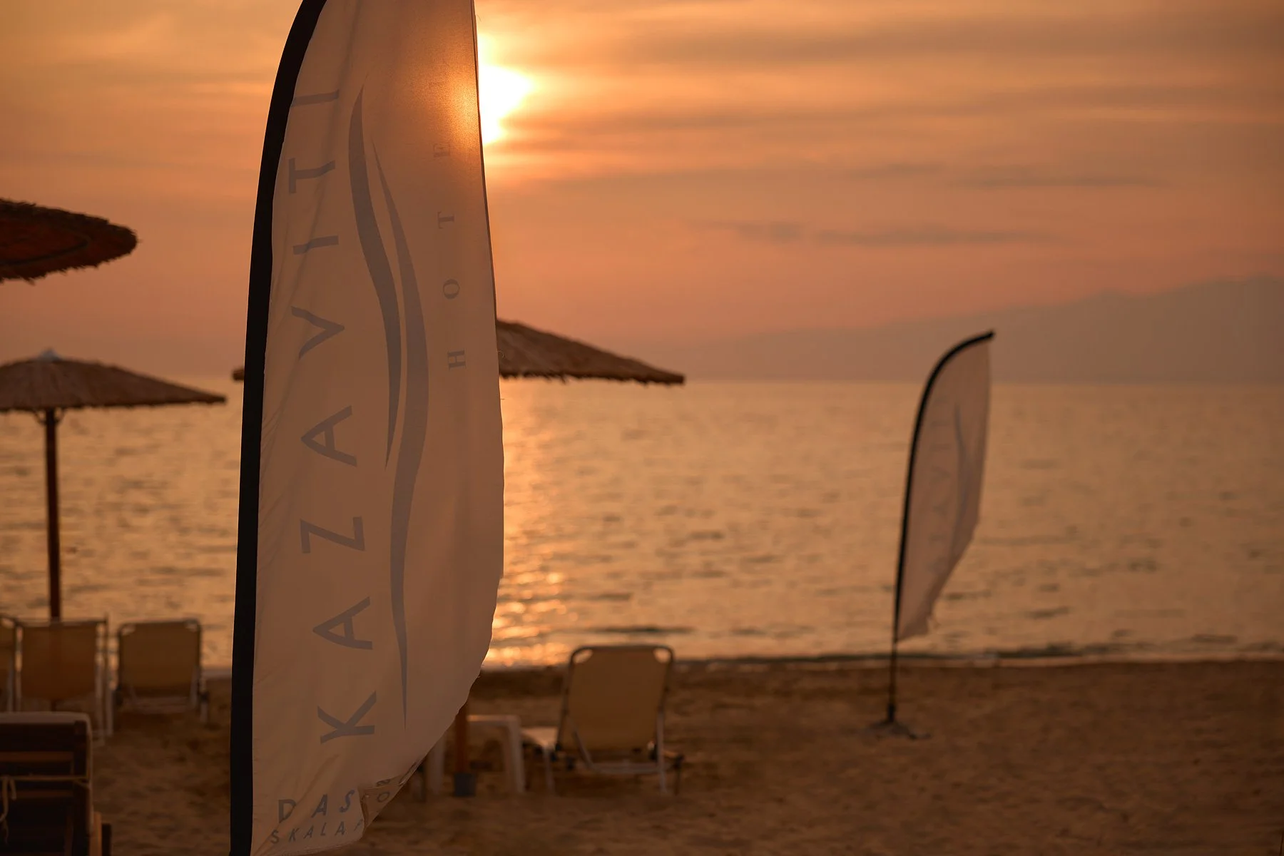 Beach scene at sunset with the sea, sandy shore, beach chairs, and umbrellas. Two flags with the text 'KALZATI' and a logo are visible.