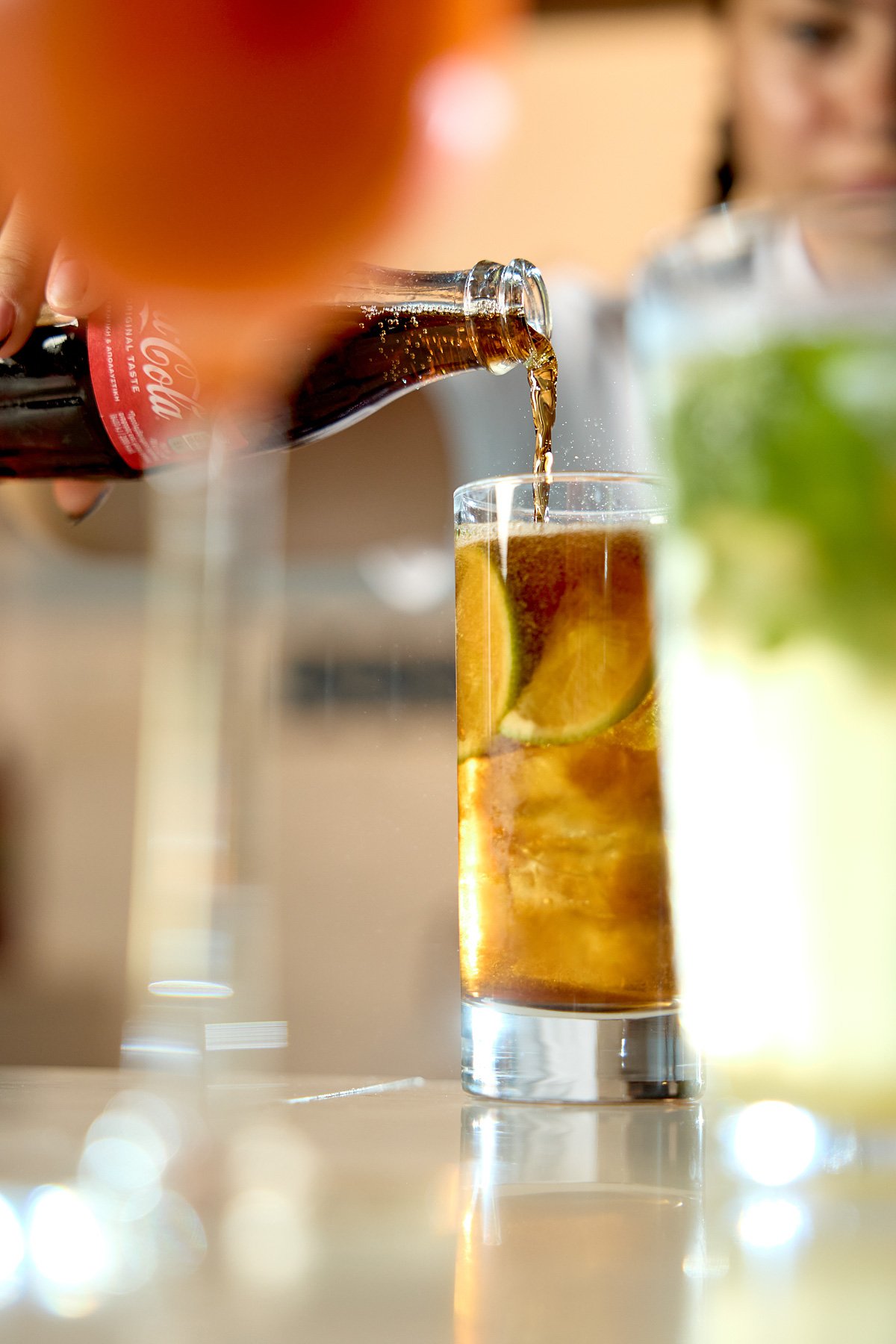 A person pours Coca-Cola from a bottle into a tall glass with lime wedges while another glass with lime and mint is visible in the background.