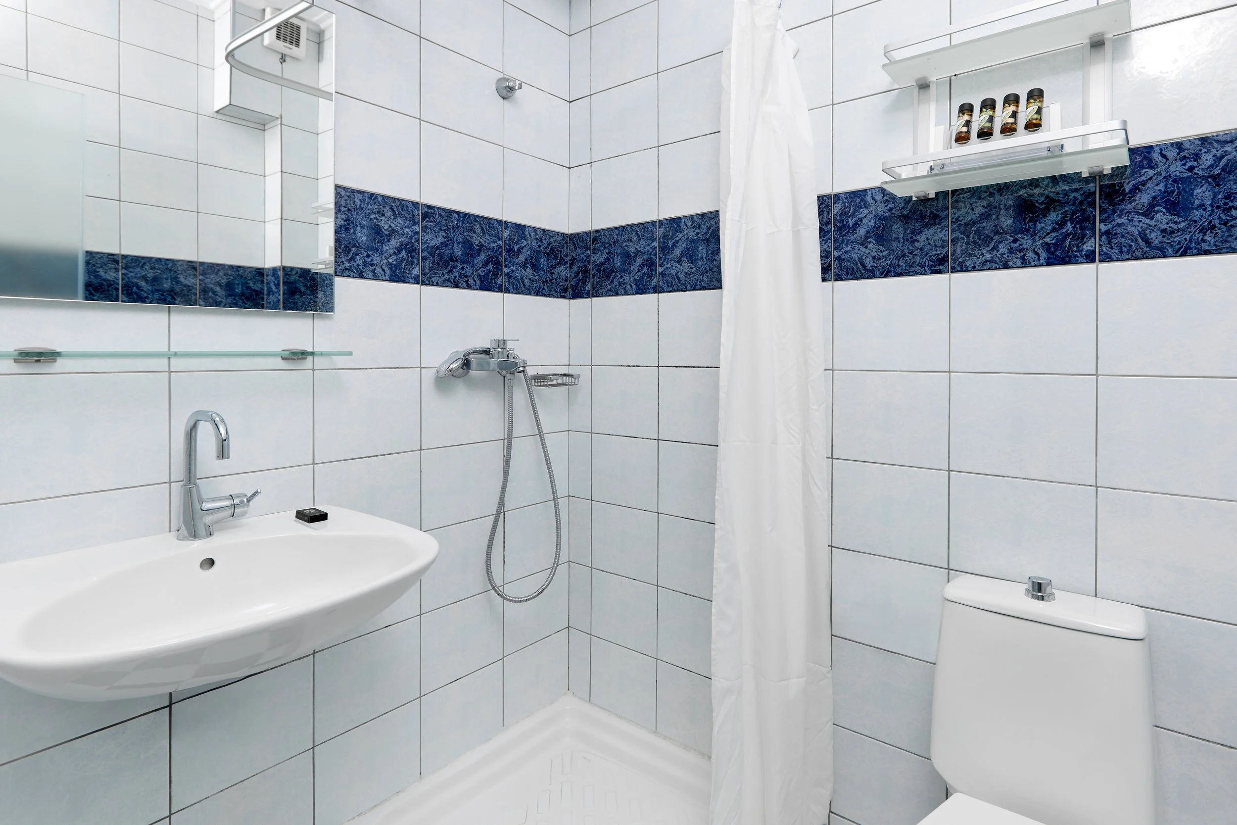 Bathroom with white tiled walls, blue accent tiles, a mirror, a small sink, a shower area with a curtain, a soap dish, and a toilet.